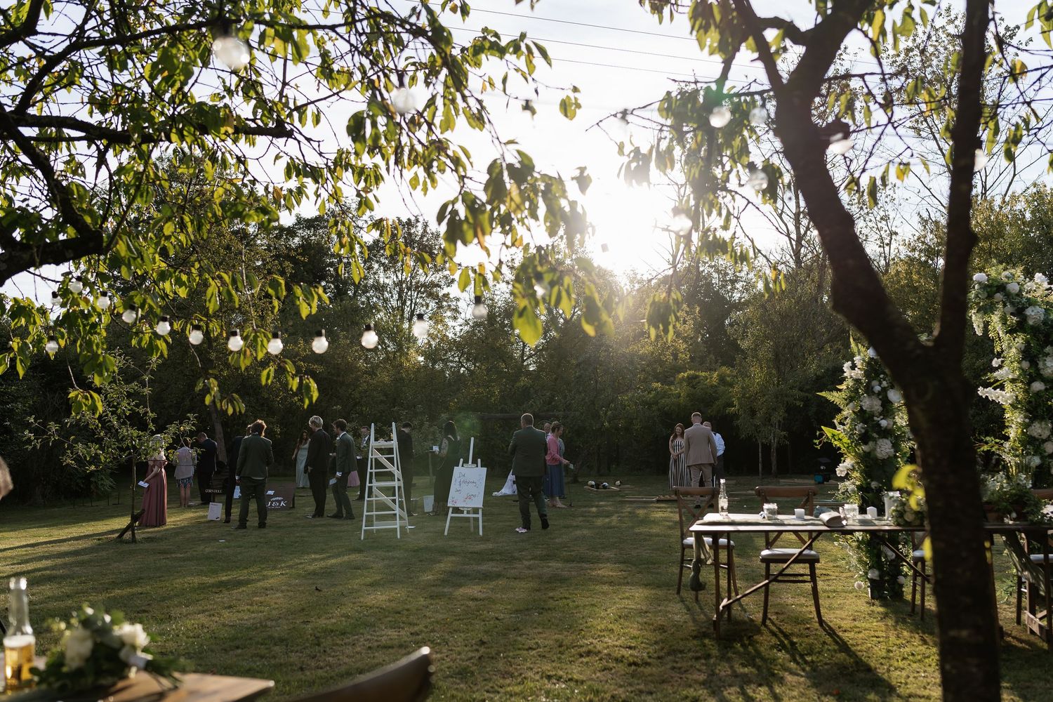 Golden hour outdoor wedding reception with guests mingling on lawn surrounded by trees and string lights.