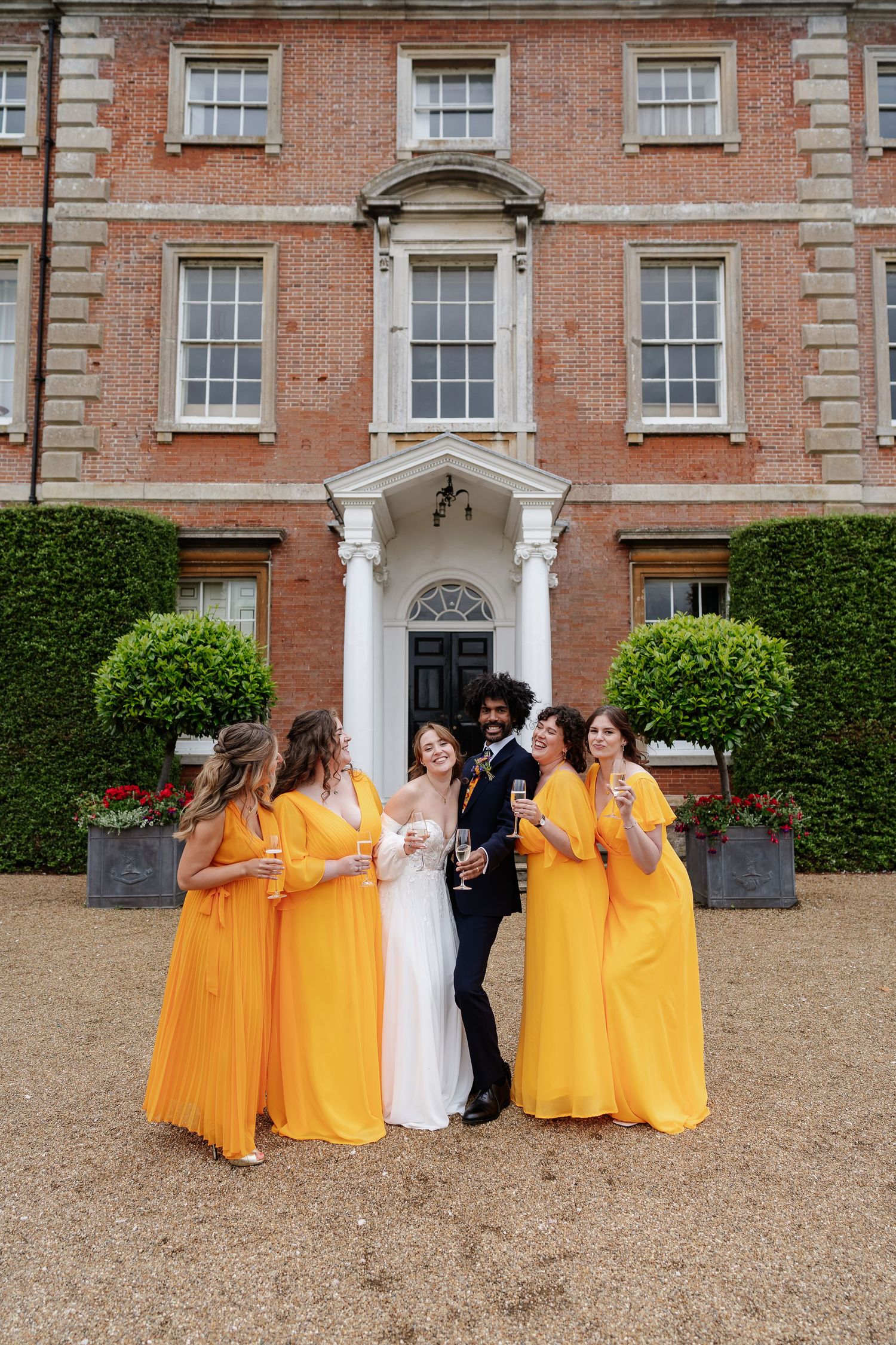 Bride and groom celebrate with four bridesmaids in matching yellow gowns outside grand brick manor.