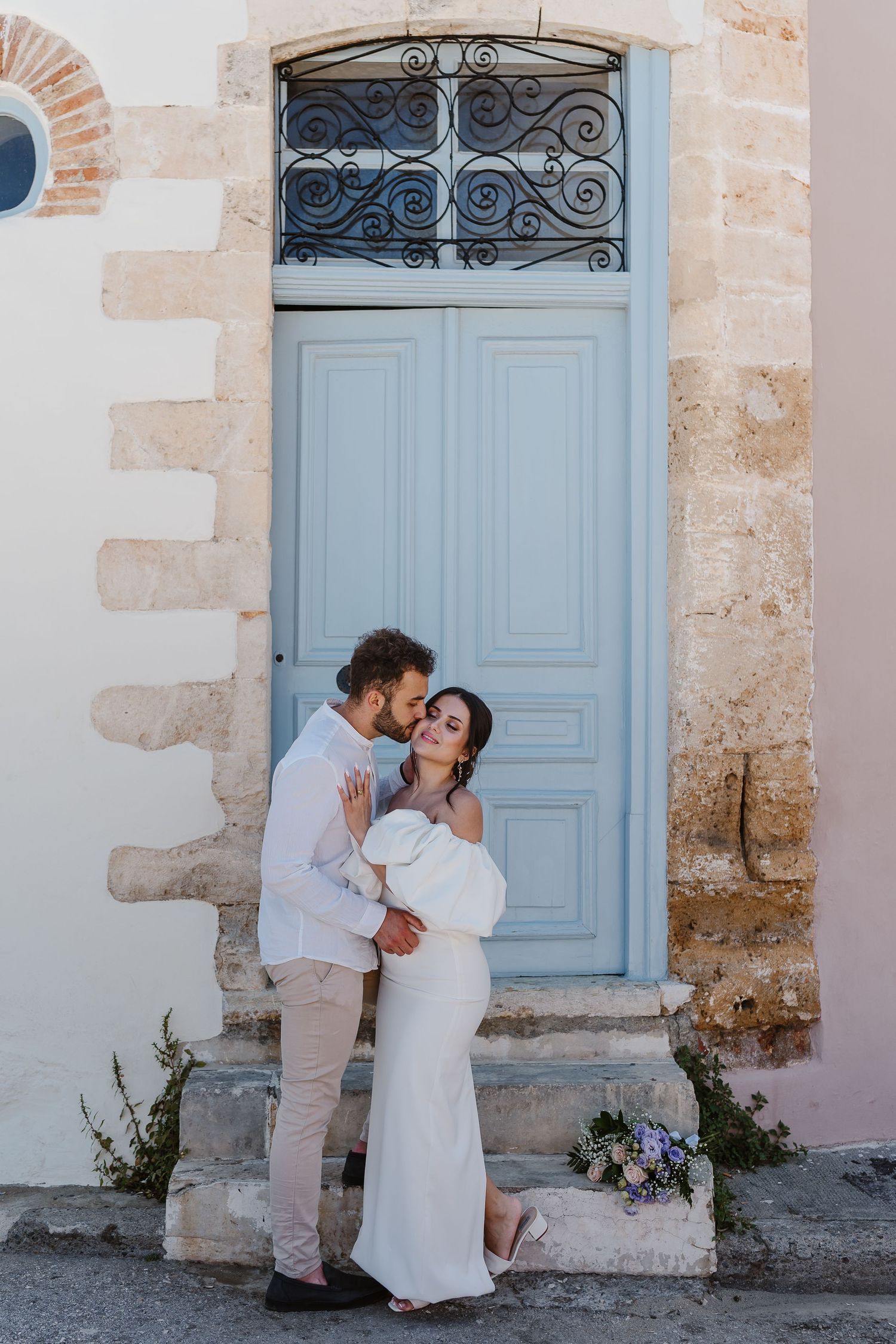 Groom tenderly kisses bride in elegant off-shoulder white gown outside a charming blue door.