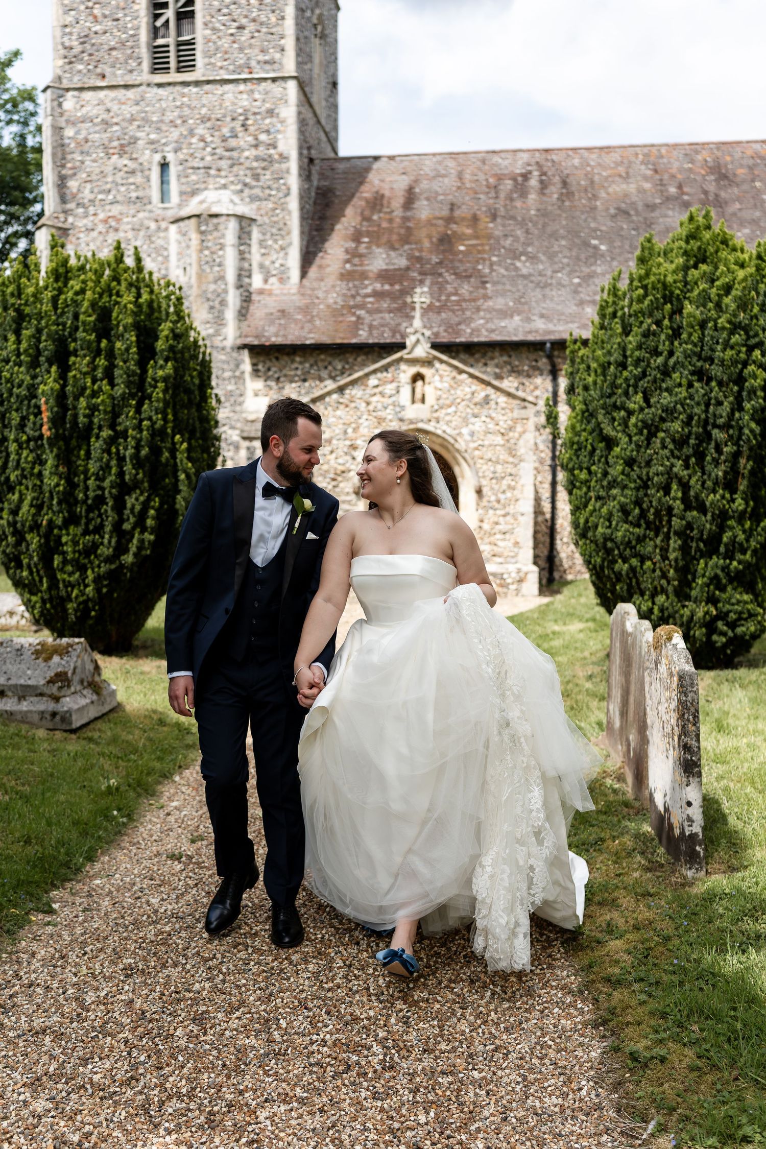 Bride and groom candidly hold hands and walk along path in front of Church on their wedding day in Suffolk.