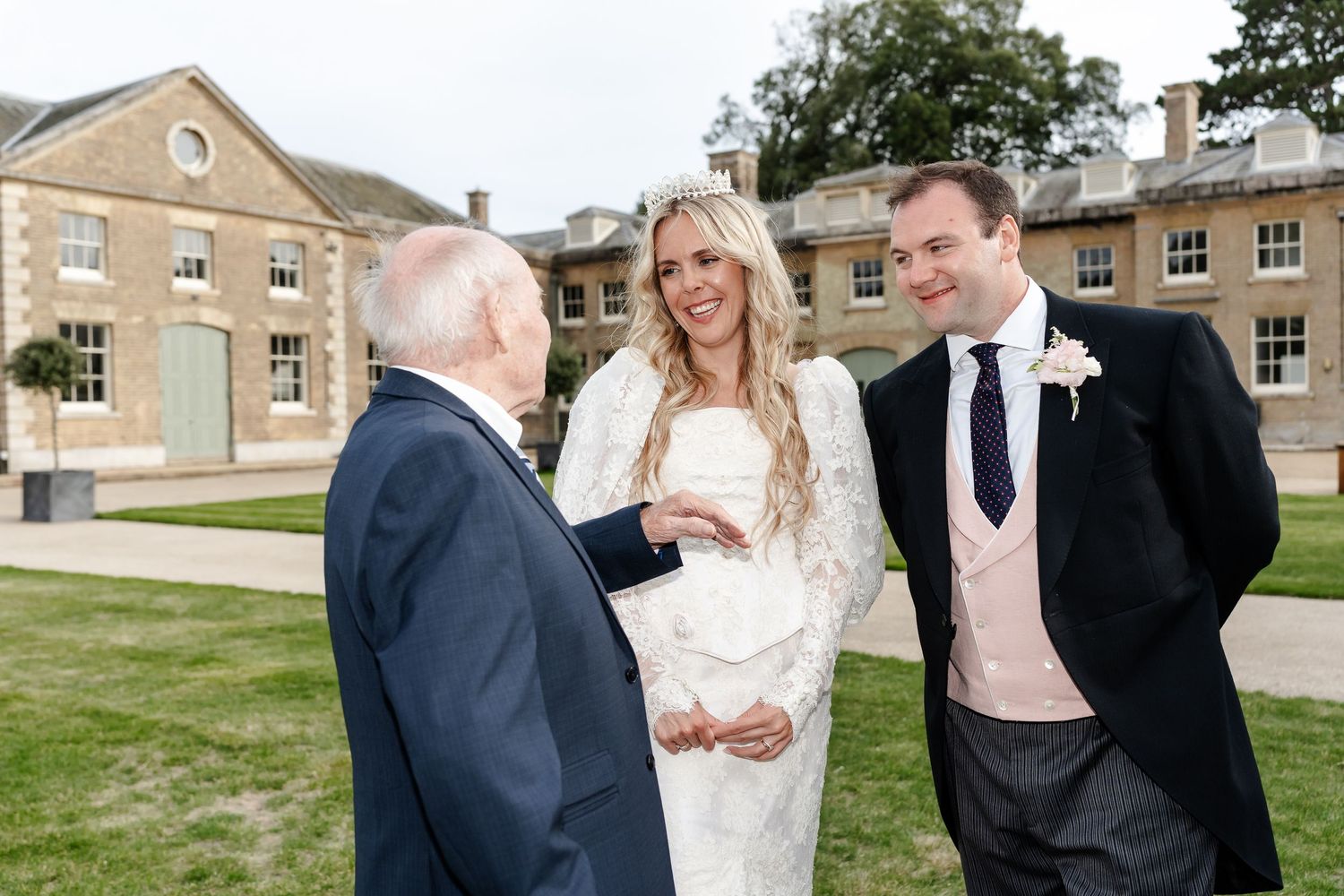 Bride in white lace dress and groom in suit chat with elderly guest outside elegant stone manor wedding venue.