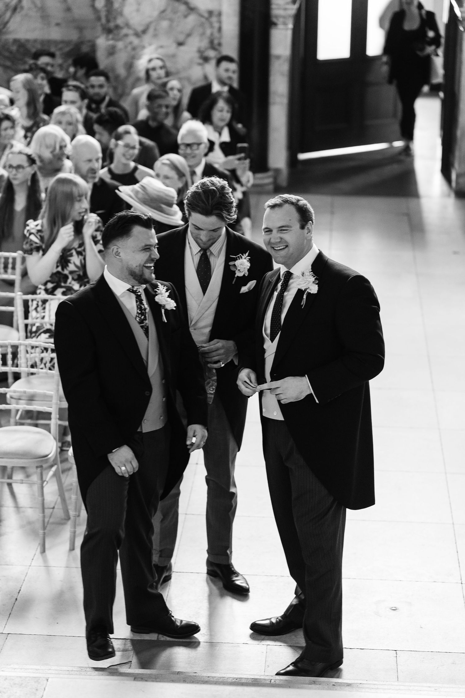 Three groomsmen laughing together at a wedding ceremony in black and white photography.