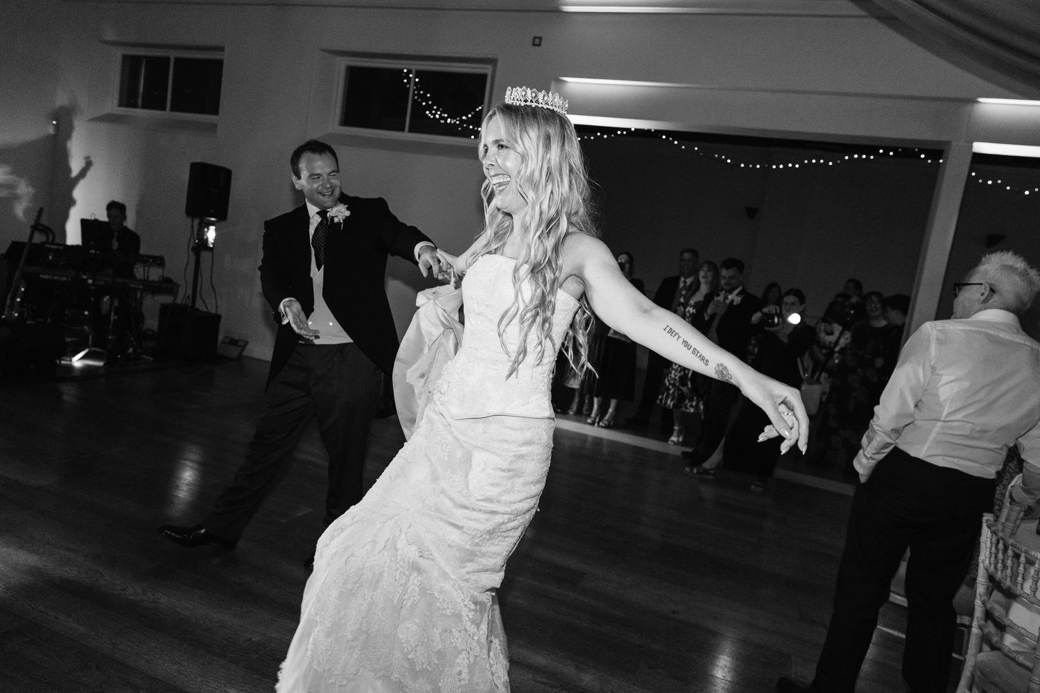 Bride dancing joyfully in a fitted lace gown during a lively black and white wedding reception.
