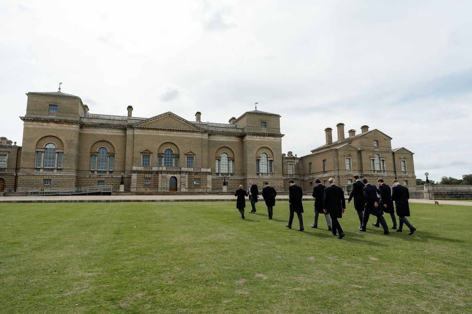 Group of people in dark suits walking across green lawn outside a grand historic stone building under cloudy skies.