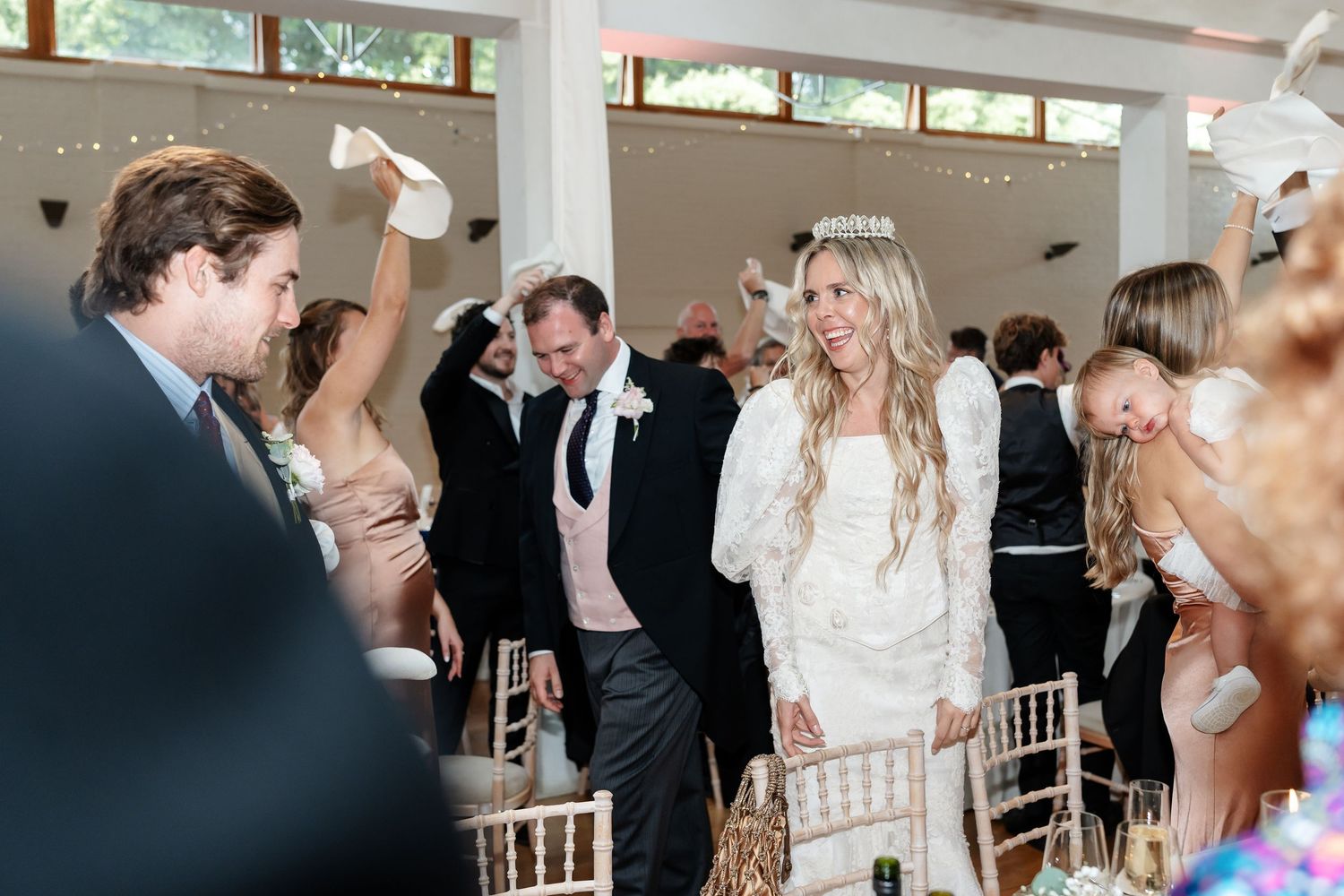 Bride in white lace gown and groom walk through cheering guests at elegant wedding reception entrance.