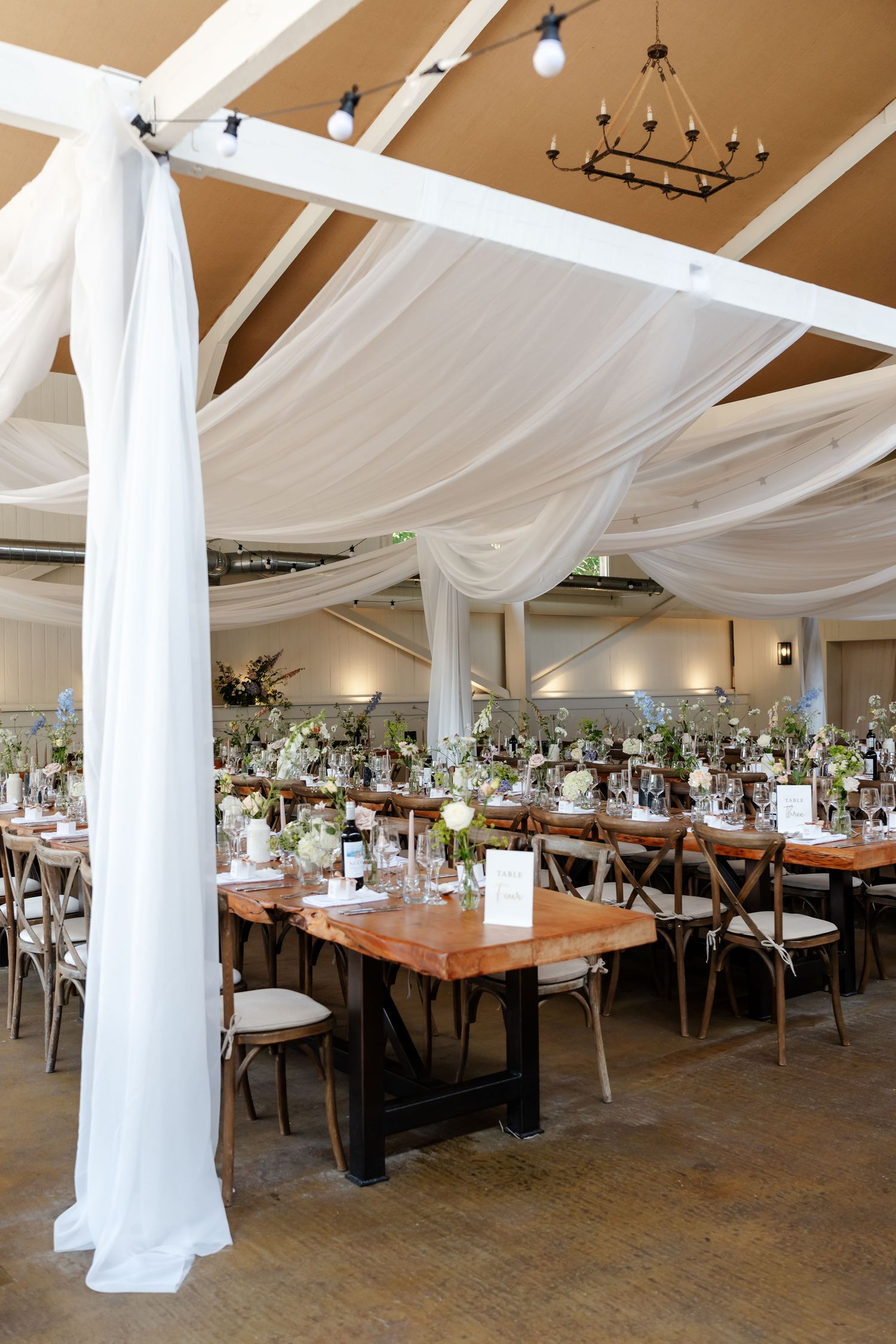 Elegant wedding reception hall with white draped ceiling, wooden farm tables, and cross-back chairs.