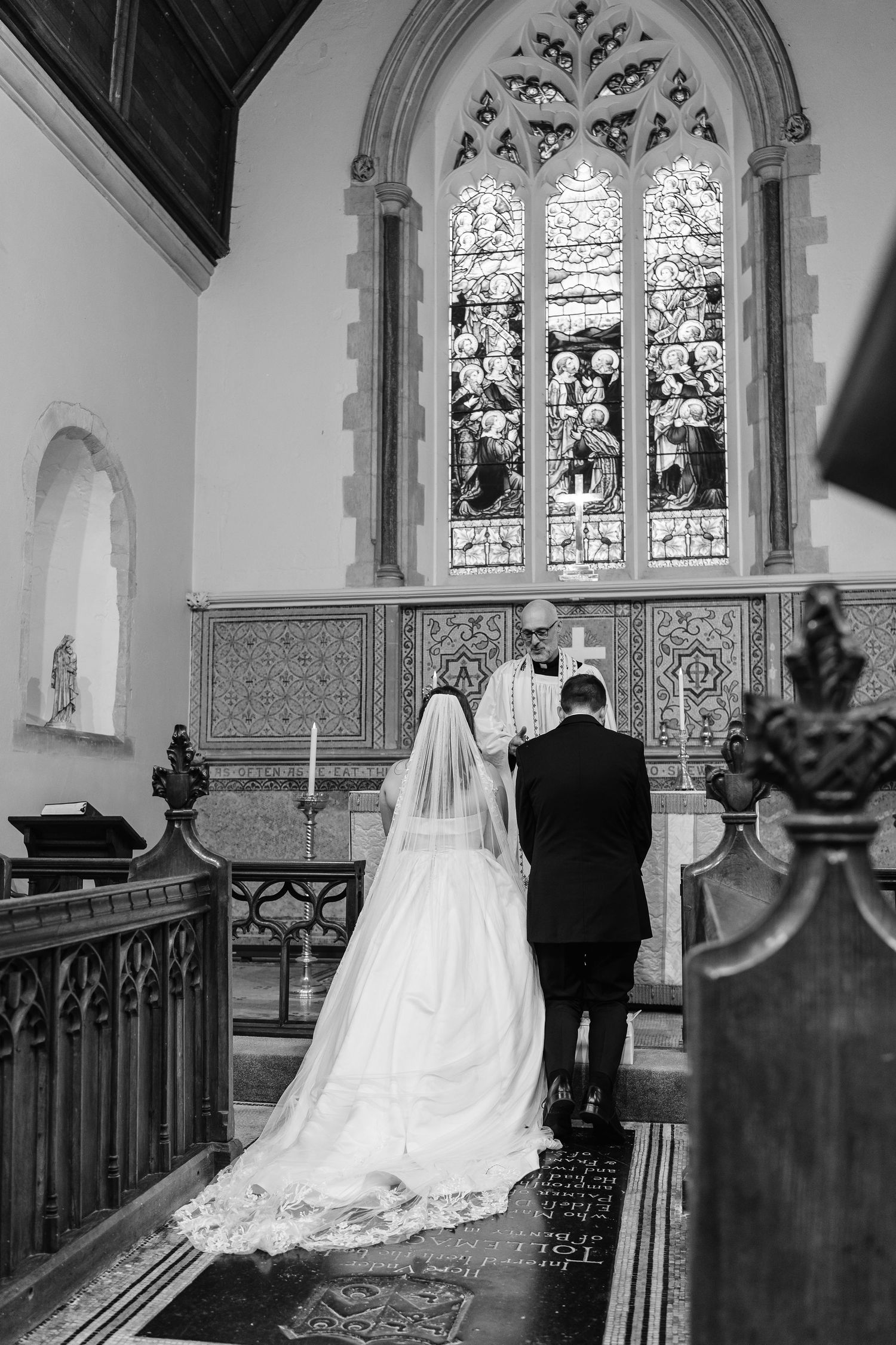 Black and white photo of bride and groom at church altar before stained glass window during ceremony.