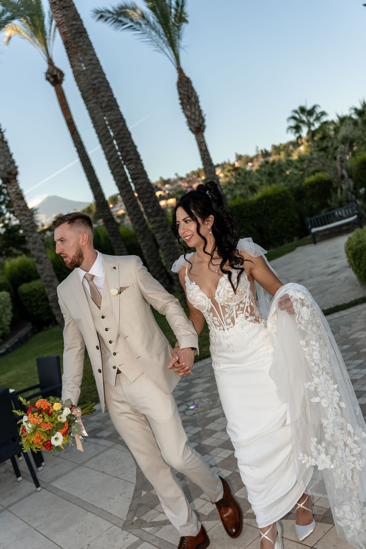 Smiling newlyweds walking hand in hand through a palm tree-lined path at sunset wedding.