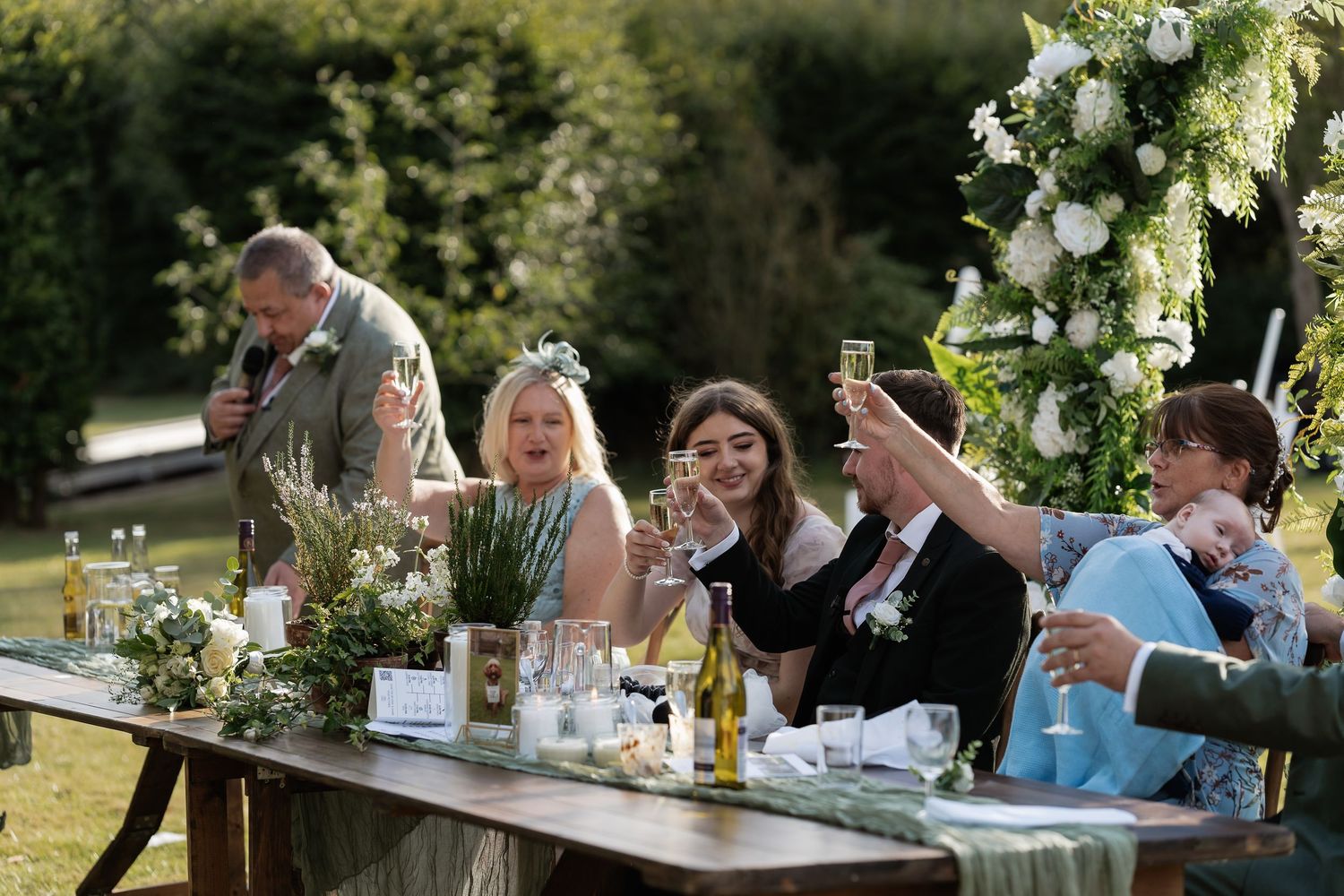 Bride and groom raise champagne glasses with guests at a floral-decorated outdoor wedding reception table.