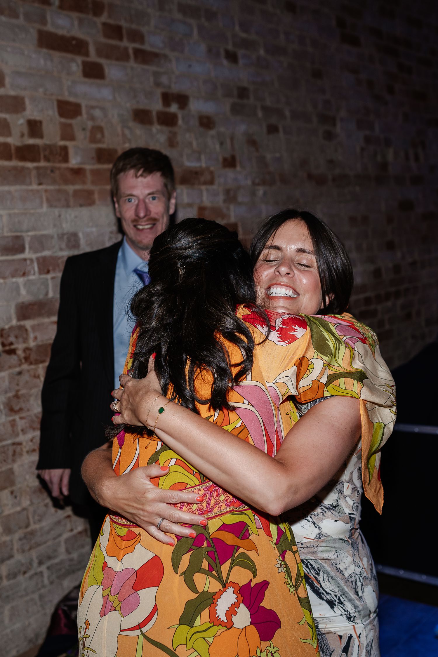 Two women share a warm emotional hug at a wedding celebration near a brick wall.
