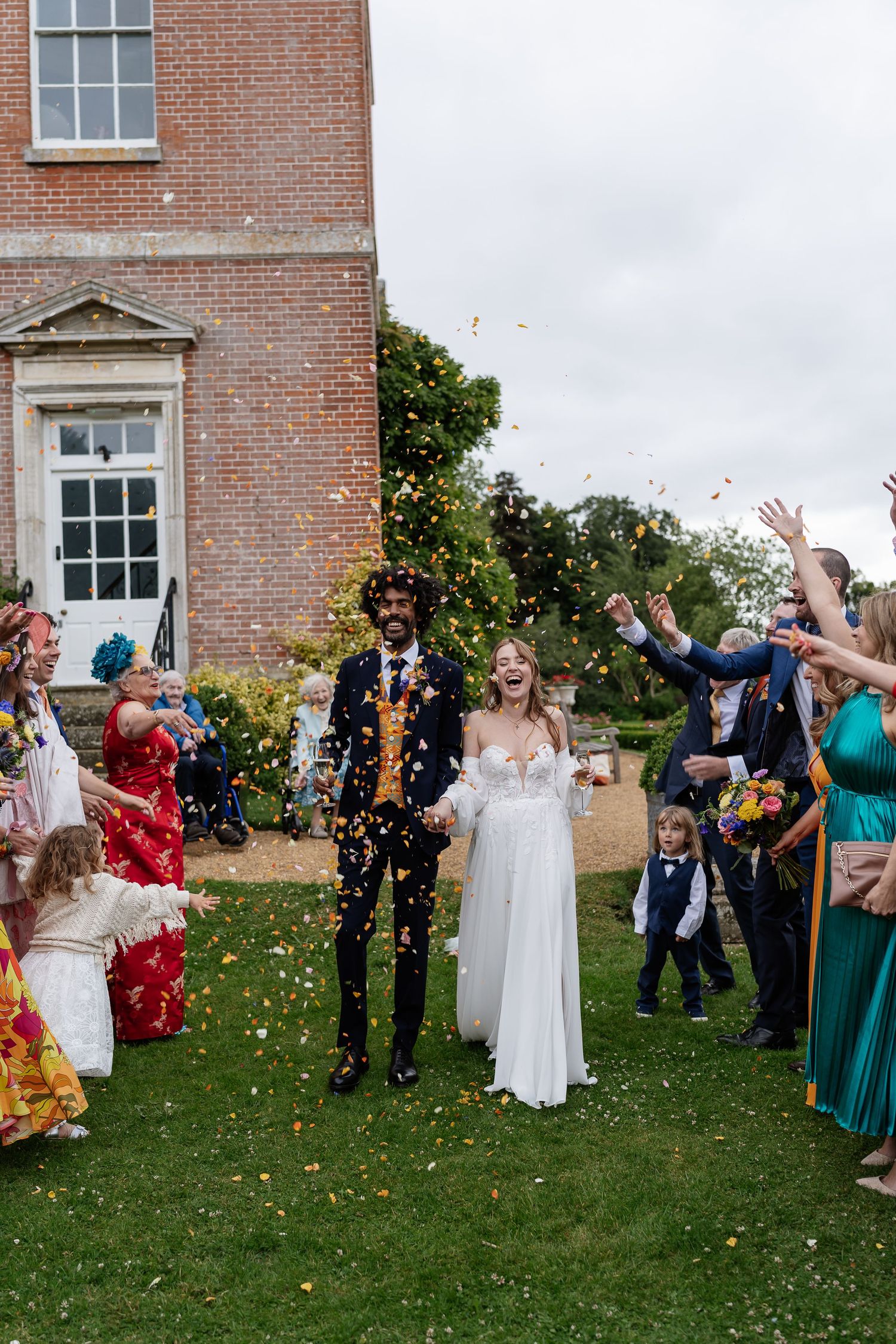 Joyful newlyweds walk through colorful confetti shower surrounded by cheering guests outside manor.