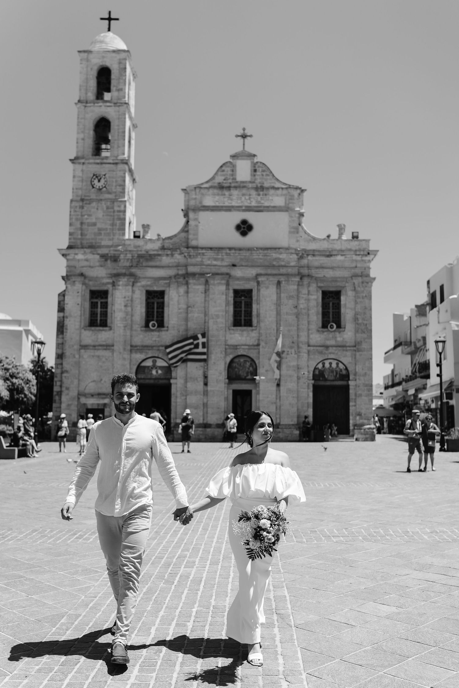 Black and white photo of bride and groom walking hand in hand in front of a historic church in Greece.