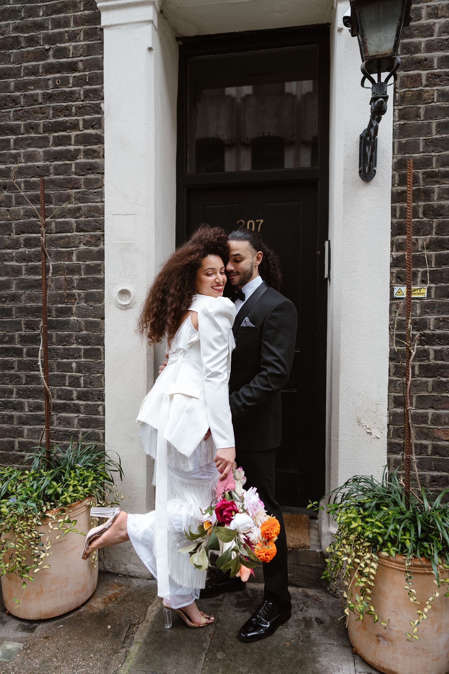 Stylish bride in white suit and groom in tuxedo embracing at elegant townhouse door.