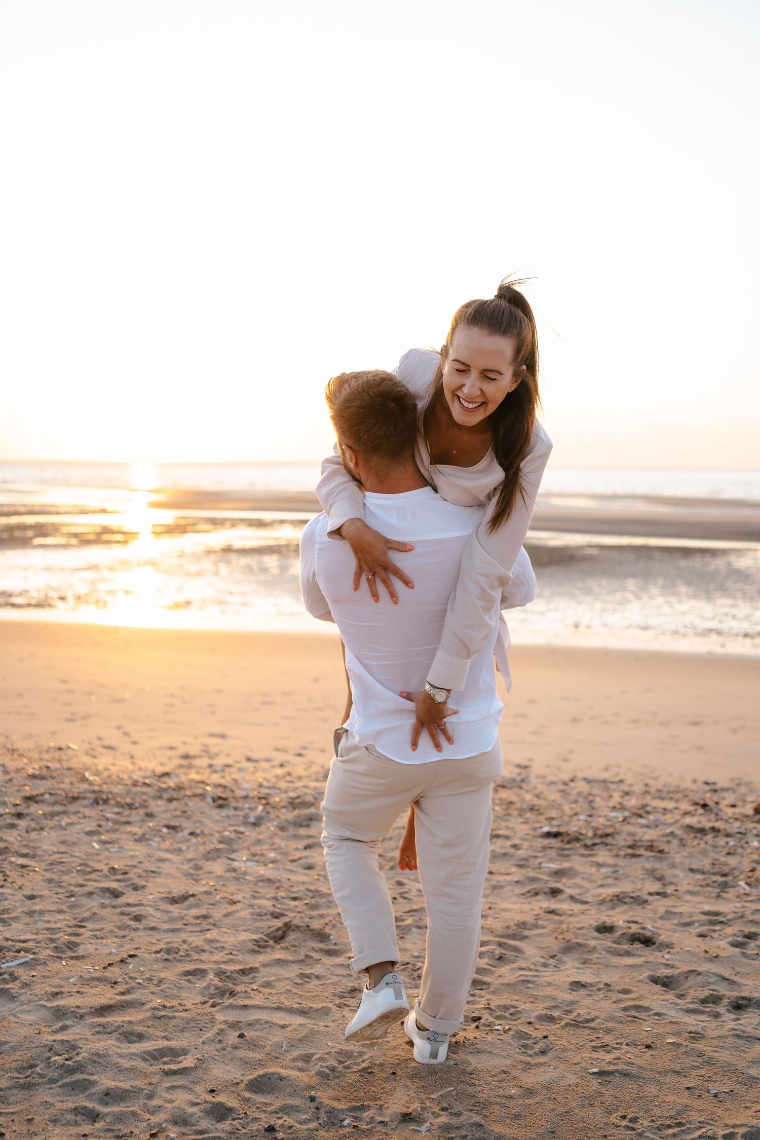 Man lifting laughing woman on sunny beach at sunset, both wearing white outfits.