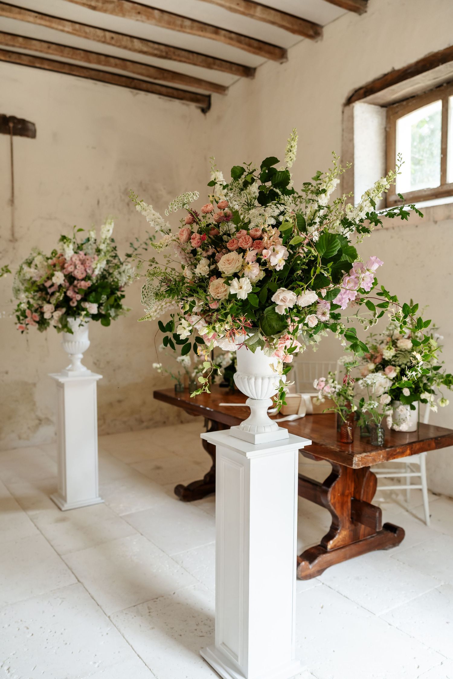 Elegant wedding ceremony decor with large floral arrangements in white urns on pedestals in a rustic venue.