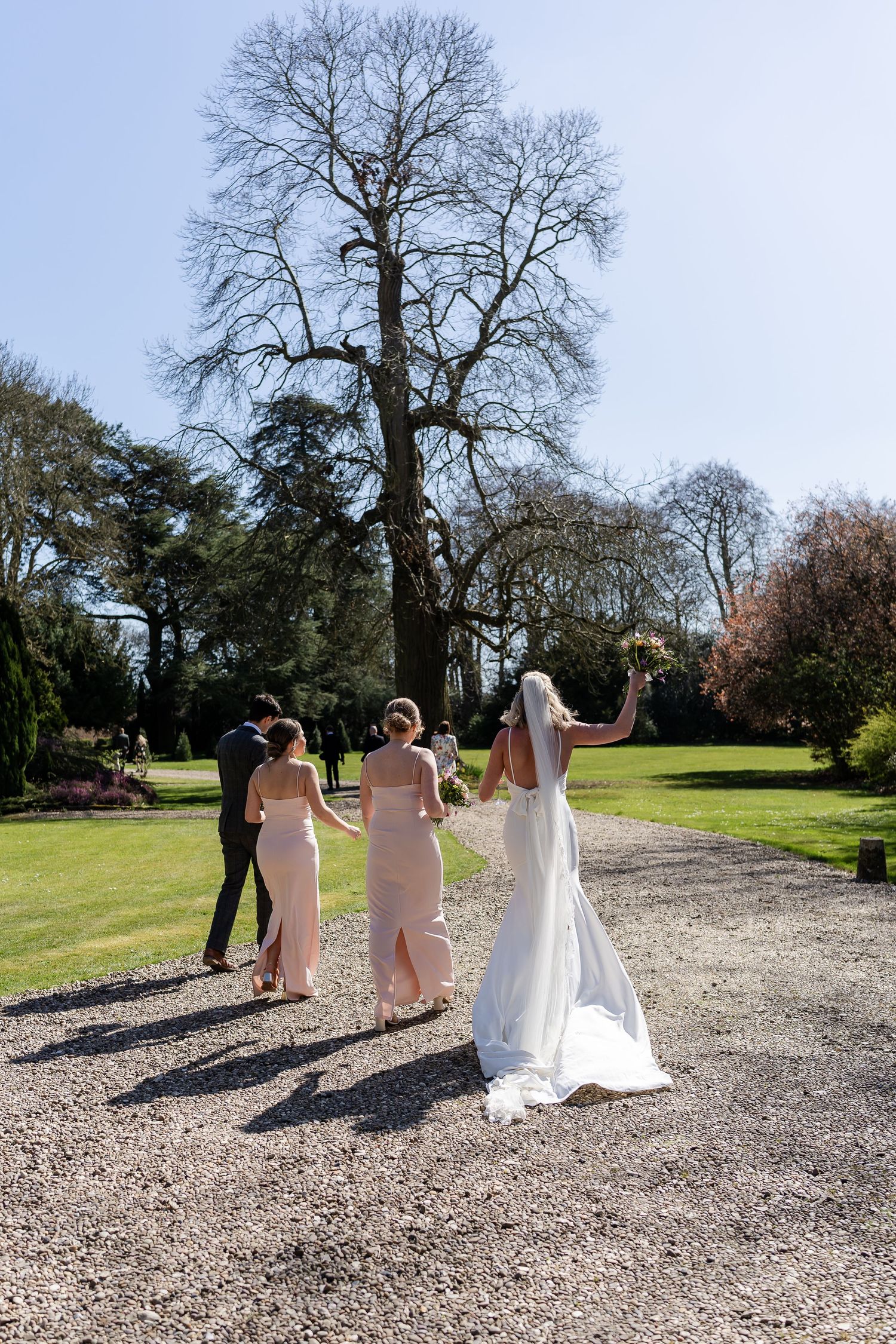 Bride in white gown raises bouquet while walking with bridesmaids in blush dresses on gravel path.