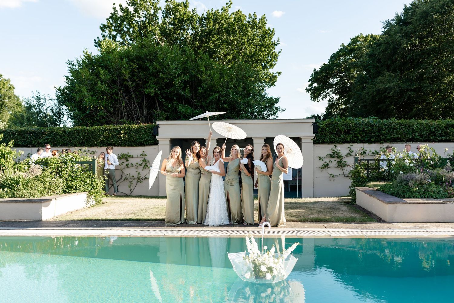 Bride poses with bridesmaids in sage green dresses beside elegant outdoor pool.