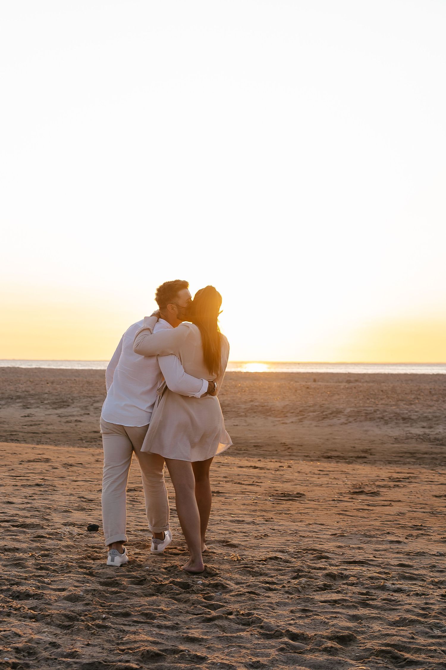 Romantic couple sharing a kiss while embracing on a sandy beach during a stunning golden sunset.