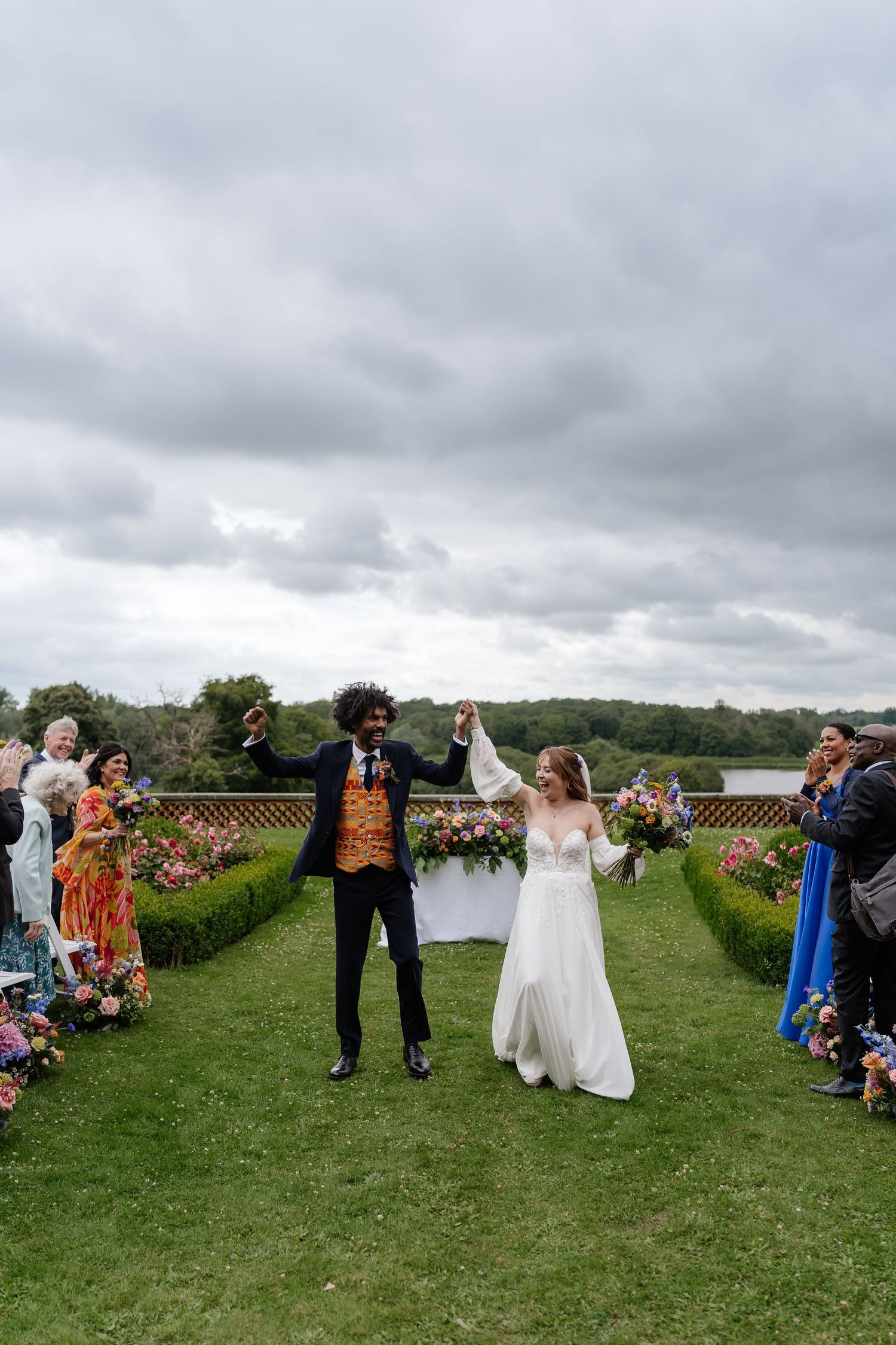 Jubilant newlyweds raise hands in celebration while walking down aisle at outdoor garden wedding ceremony.