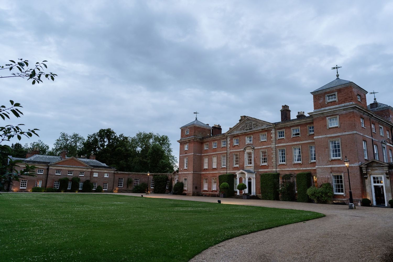 A grand Georgian red brick manor house with manicured lawns under a cloudy sky.