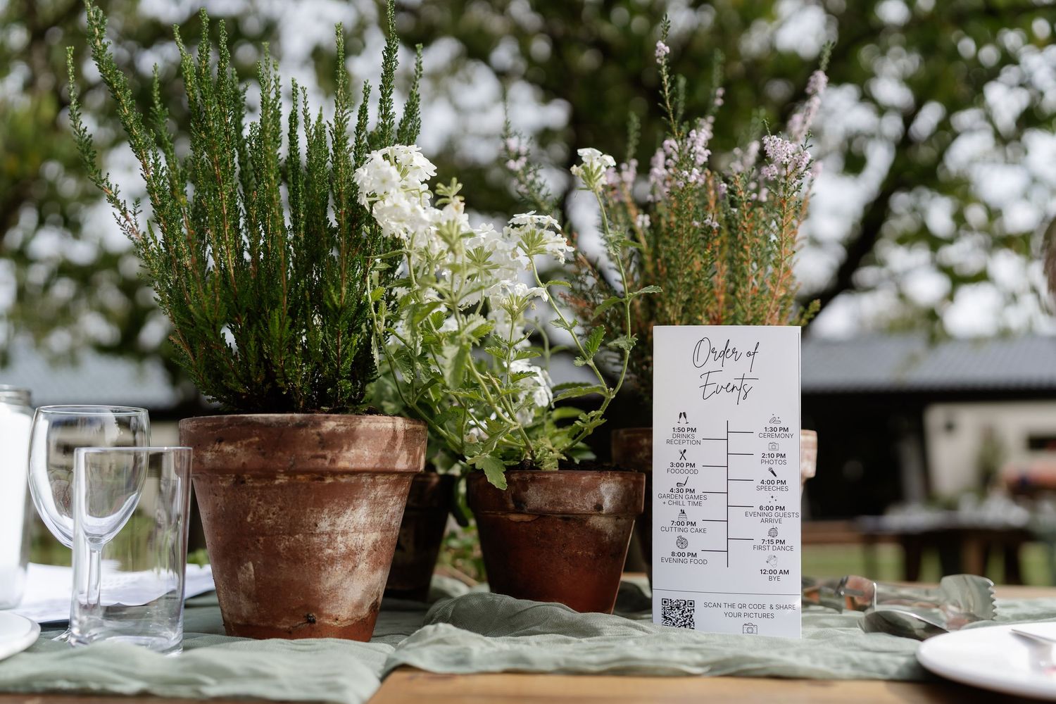 Rustic terracotta pots with herbs and white flowers beside an order of events card at outdoor wedding.
