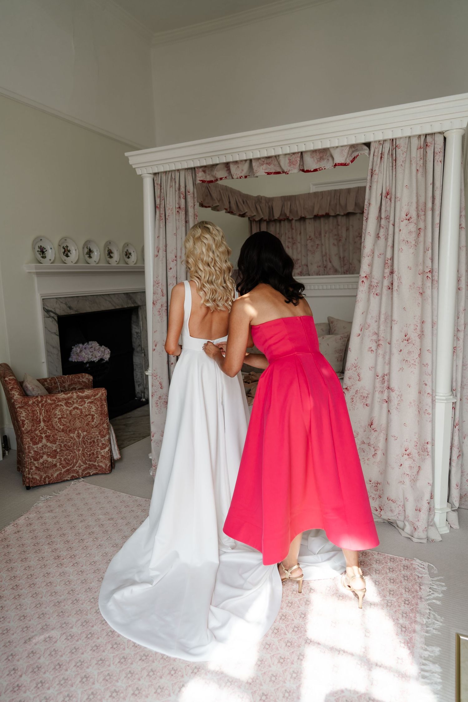 Bridesmaid in red dress helping bride with her white gown in an elegant bridal suite.