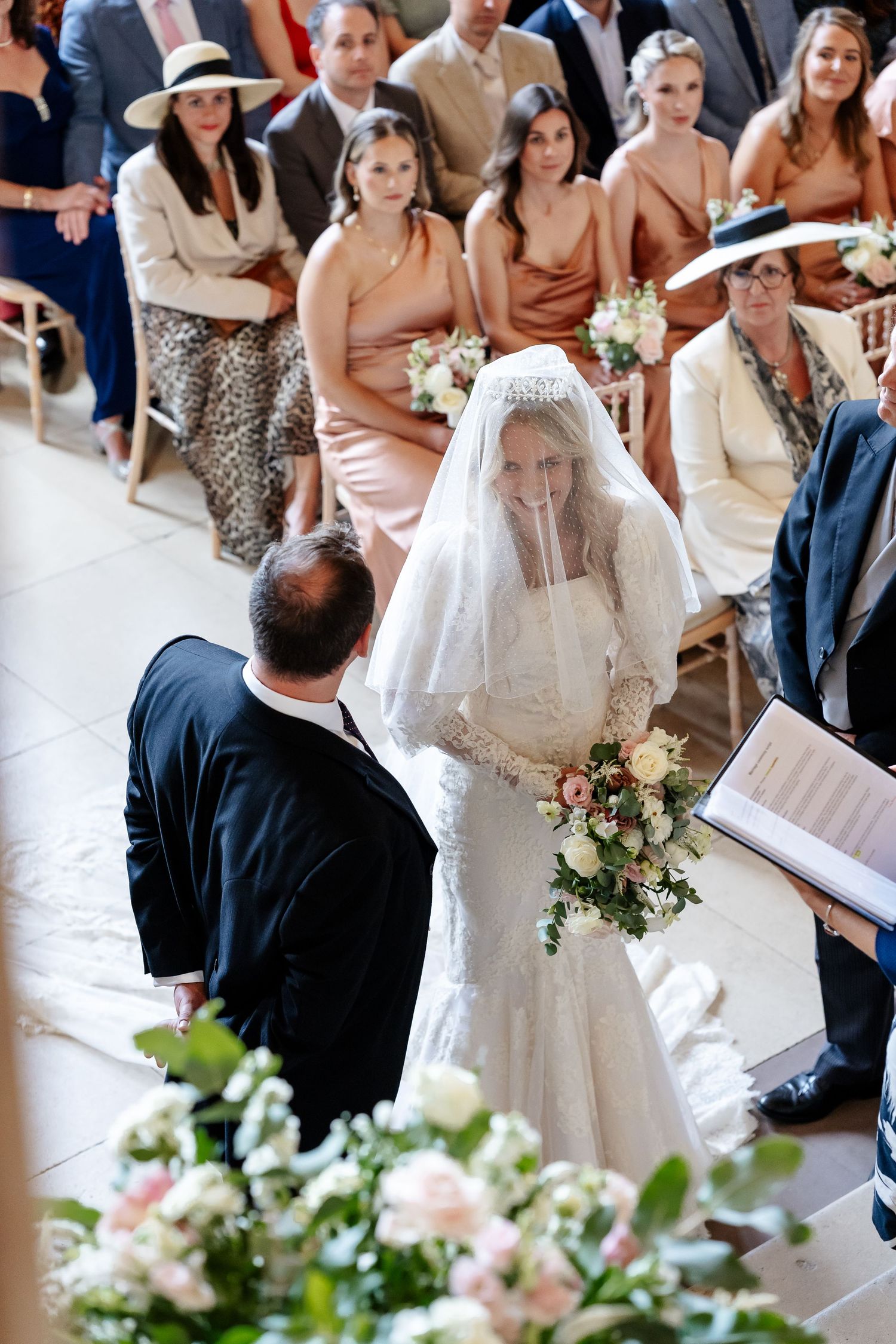 Emotional bride in lace gown and veil holds bouquet at wedding ceremony altar.
