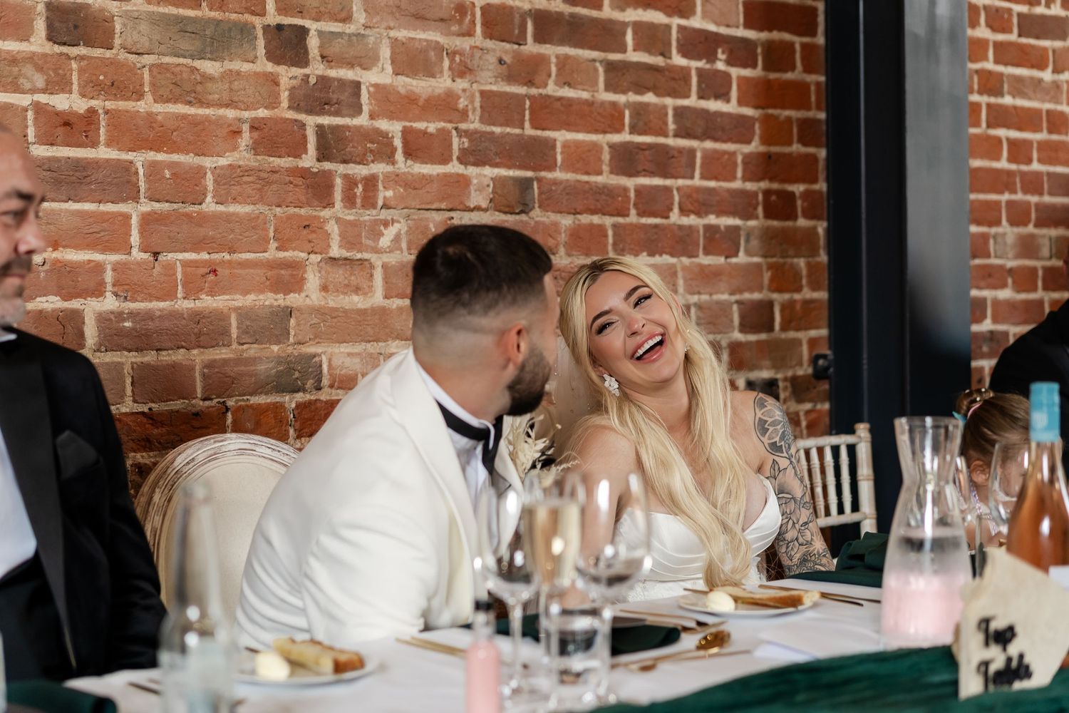 Bride laughs joyfully with groom at wedding reception head table against exposed brick wall with elegant glassware.