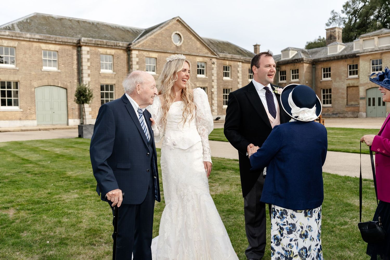 Bride and groom greet elderly guests on lawn outside elegant country house venue after wedding ceremony.