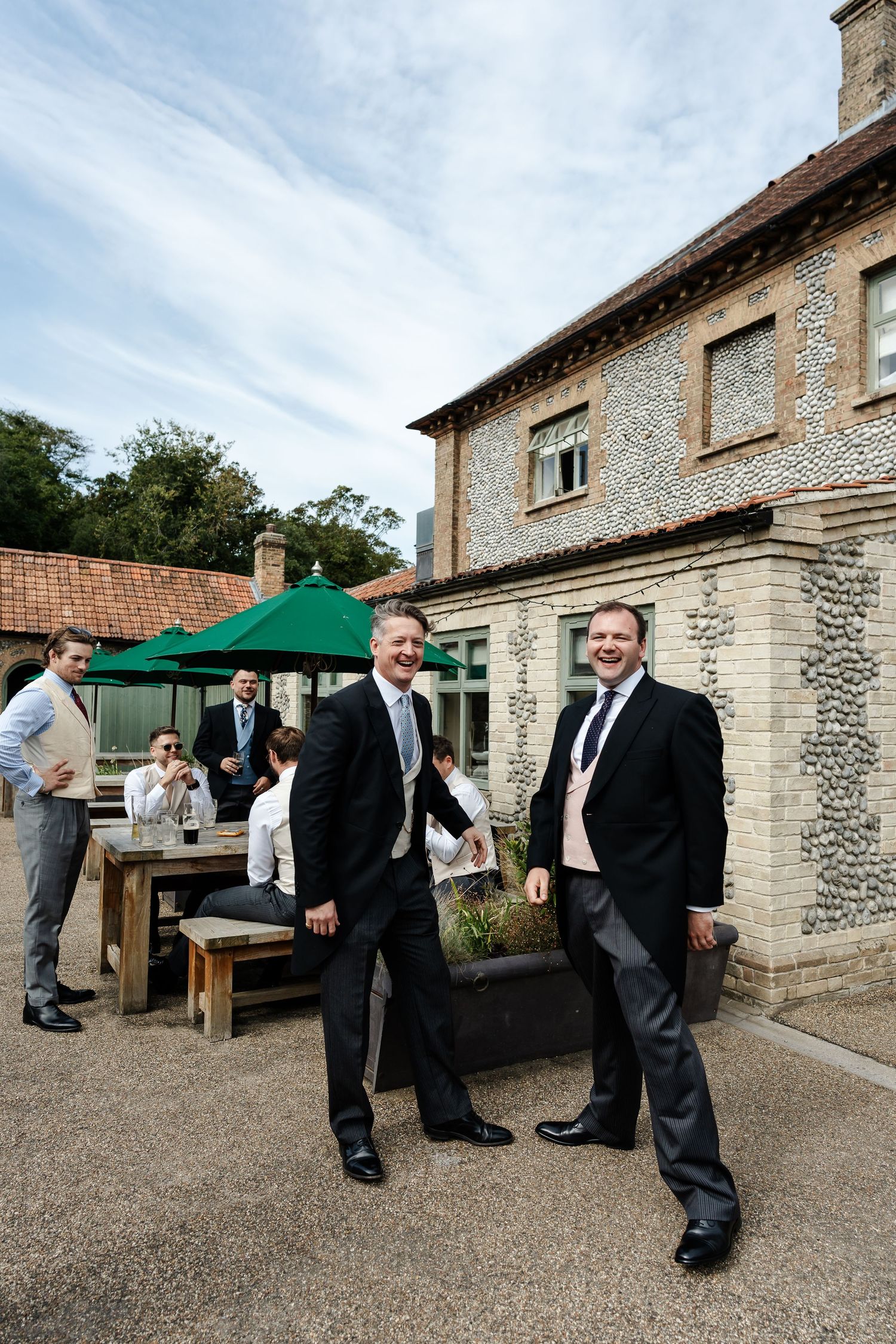 Two men in dark suits smile outdoors at a wedding venue, with guests socializing near a stone building.