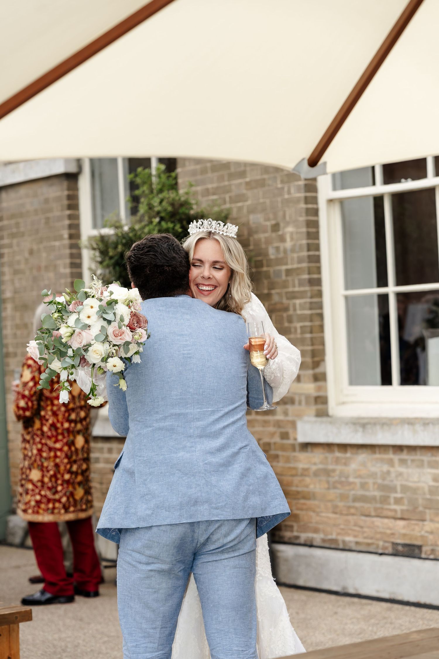 Smiling bride wearing tiara hugs groom in blue suit outdoors at wedding venue, holding champagne and bouquet.