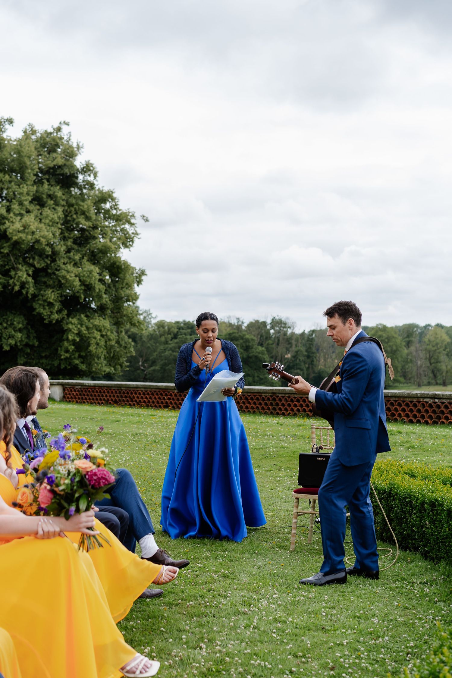 Wedding guest in blue gown reads speech while guitarist performs during outdoor garden wedding ceremony.