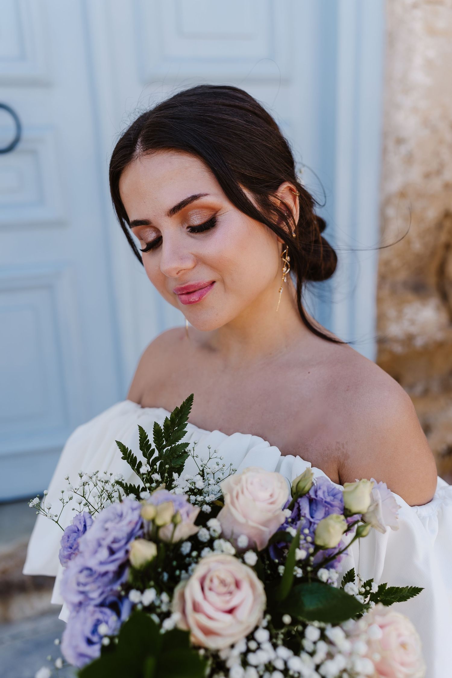 Elegant bride with updo hairstyle gazes down at a pastel bouquet of roses and lavender flowers.