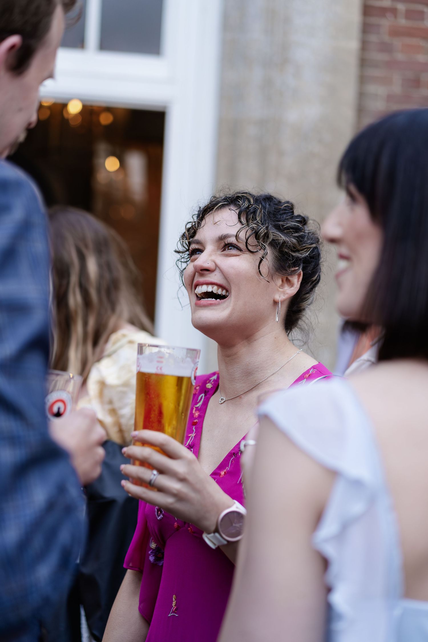 A laughing woman in a pink dress holds a beer at an outdoor wedding reception party.