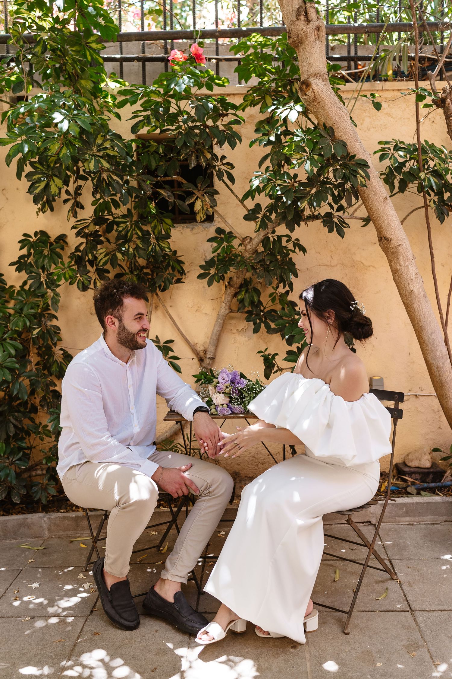 Bride and groom holding hands while seated in a sunlit courtyard garden with flowering trees.