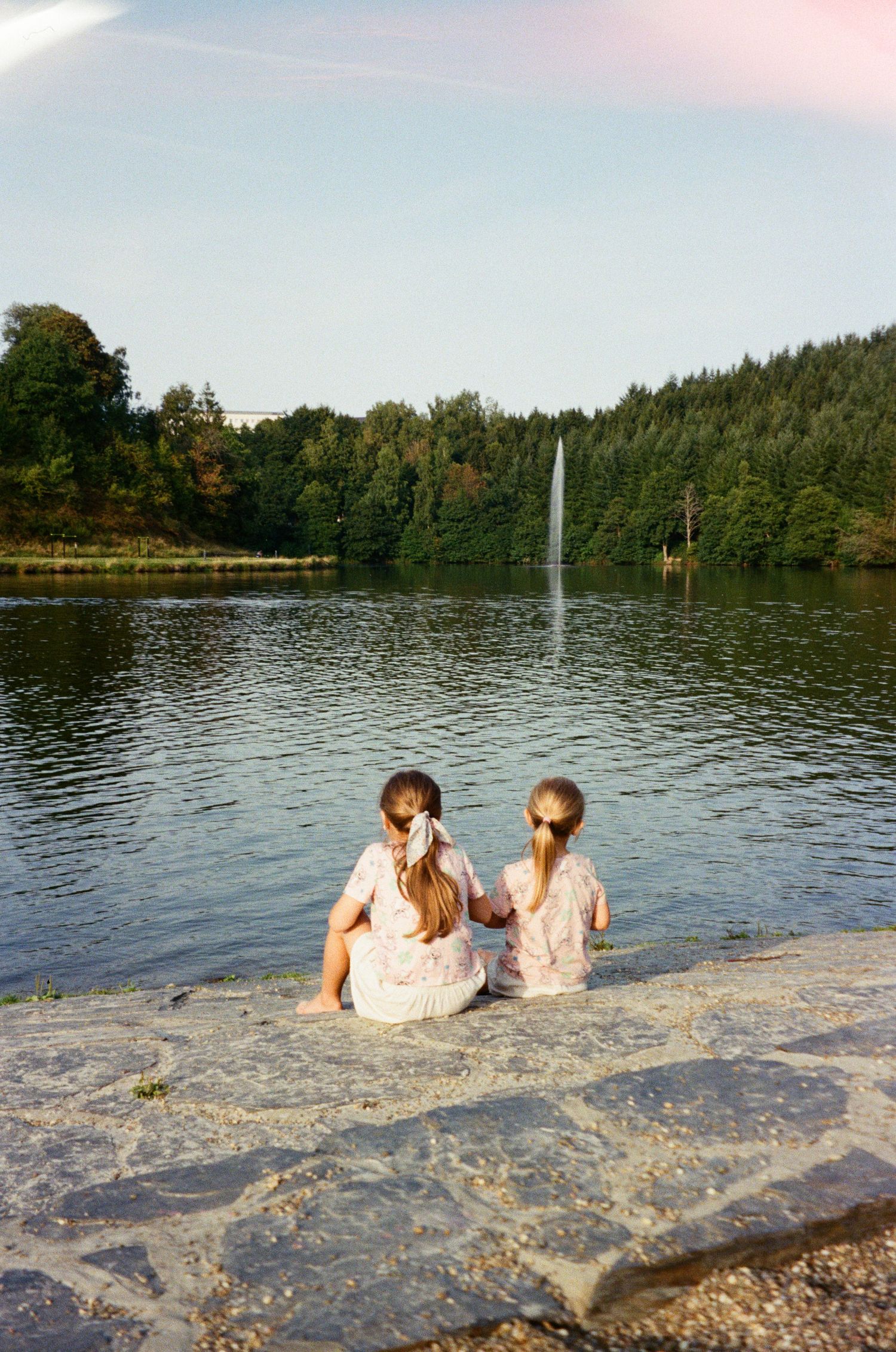 Two girls sitting by a serene lake with a fountain and lush green trees in the background.