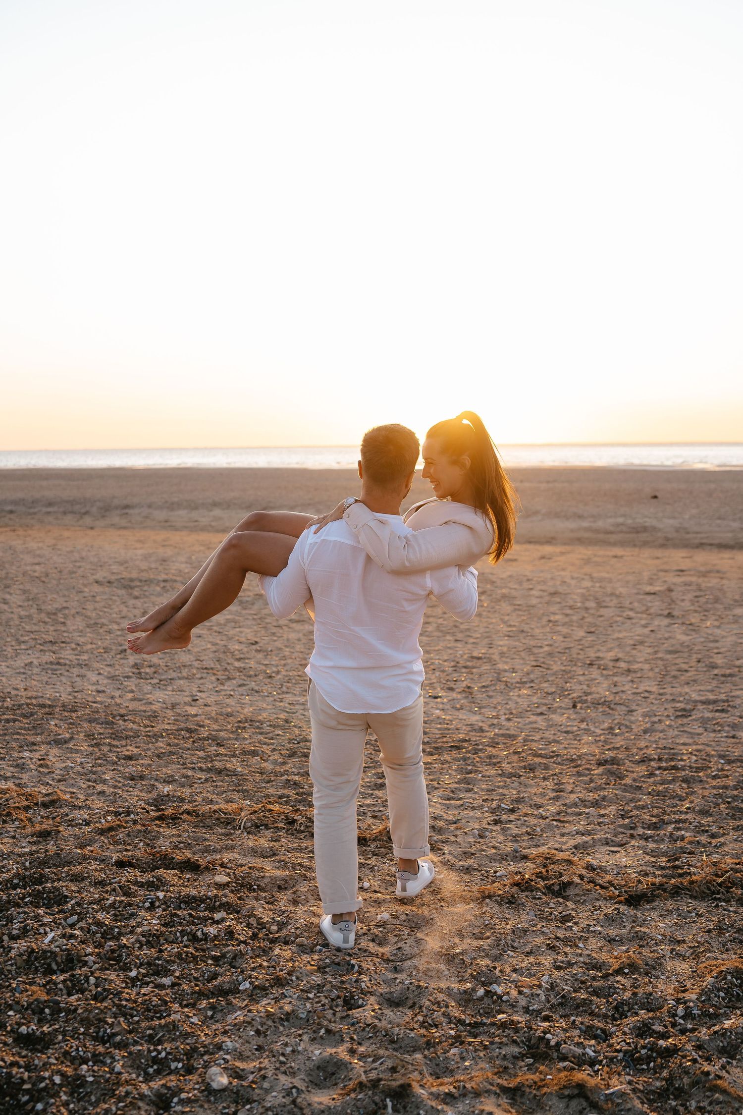 Man in white outfit carrying woman in his arms on a sandy beach during a golden sunset.