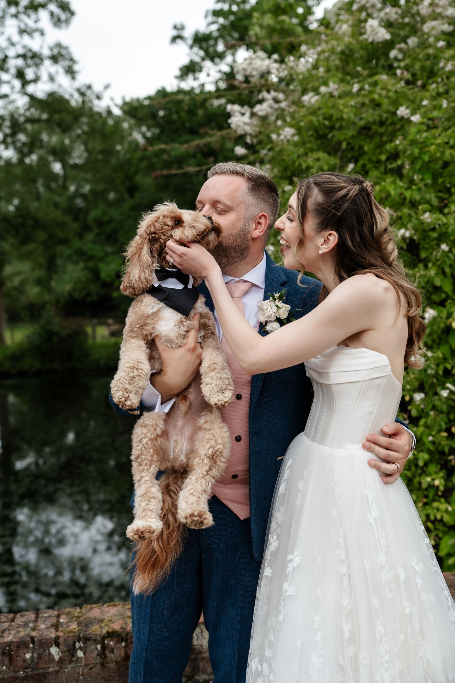 Bride and groom kissing their cute curly dog in a tuxedo at an outdoor wedding venue.