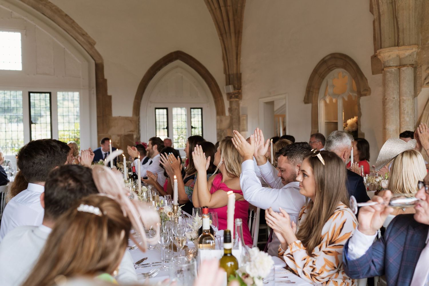 Wedding guests raise hands and cheer around a long banquet table in a stone hall at Suffolks luxury wedding venue Butley Priory