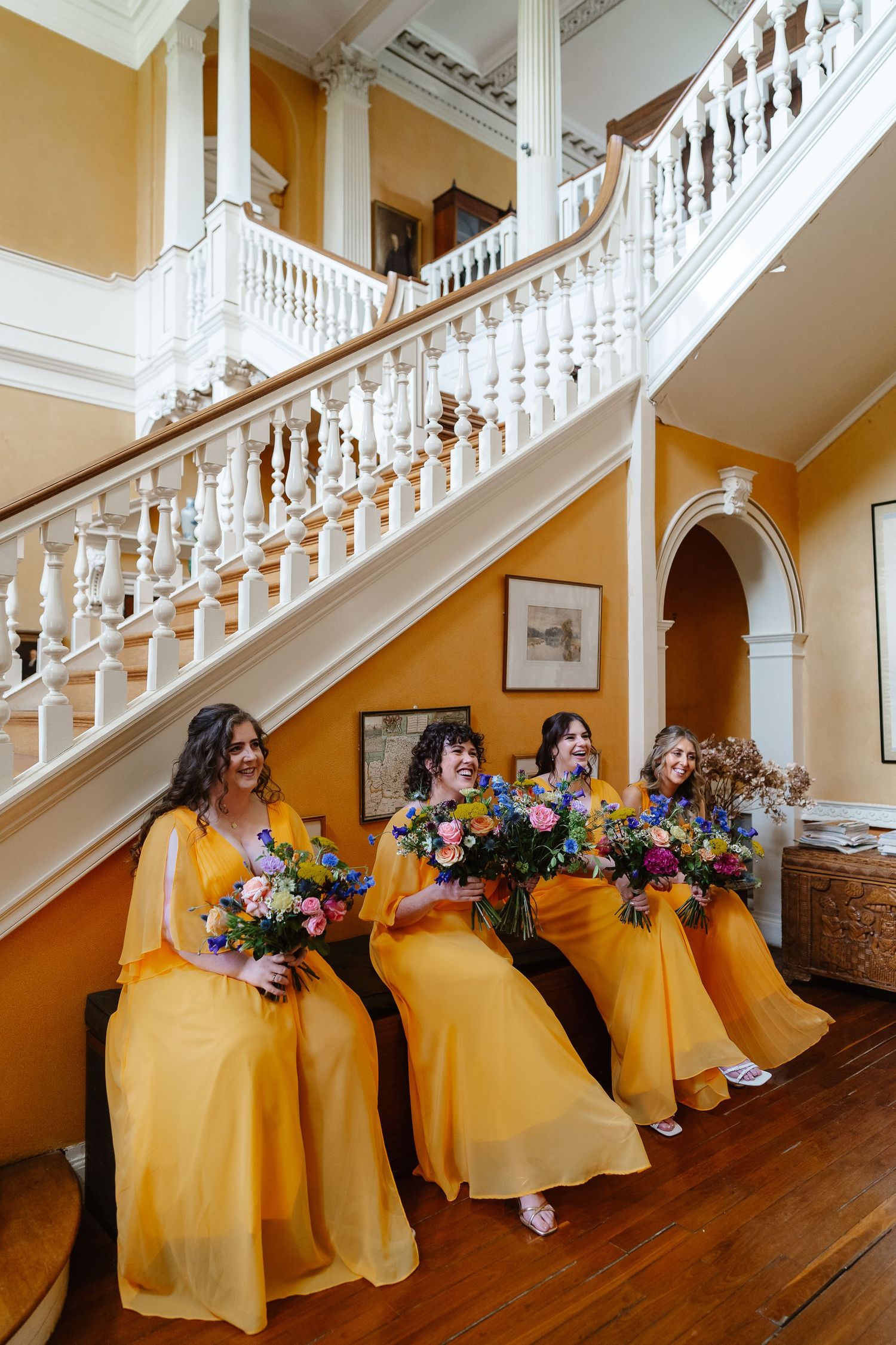 Three bridesmaids in vibrant yellow gowns holding colorful bouquets laughing on a grand staircase.