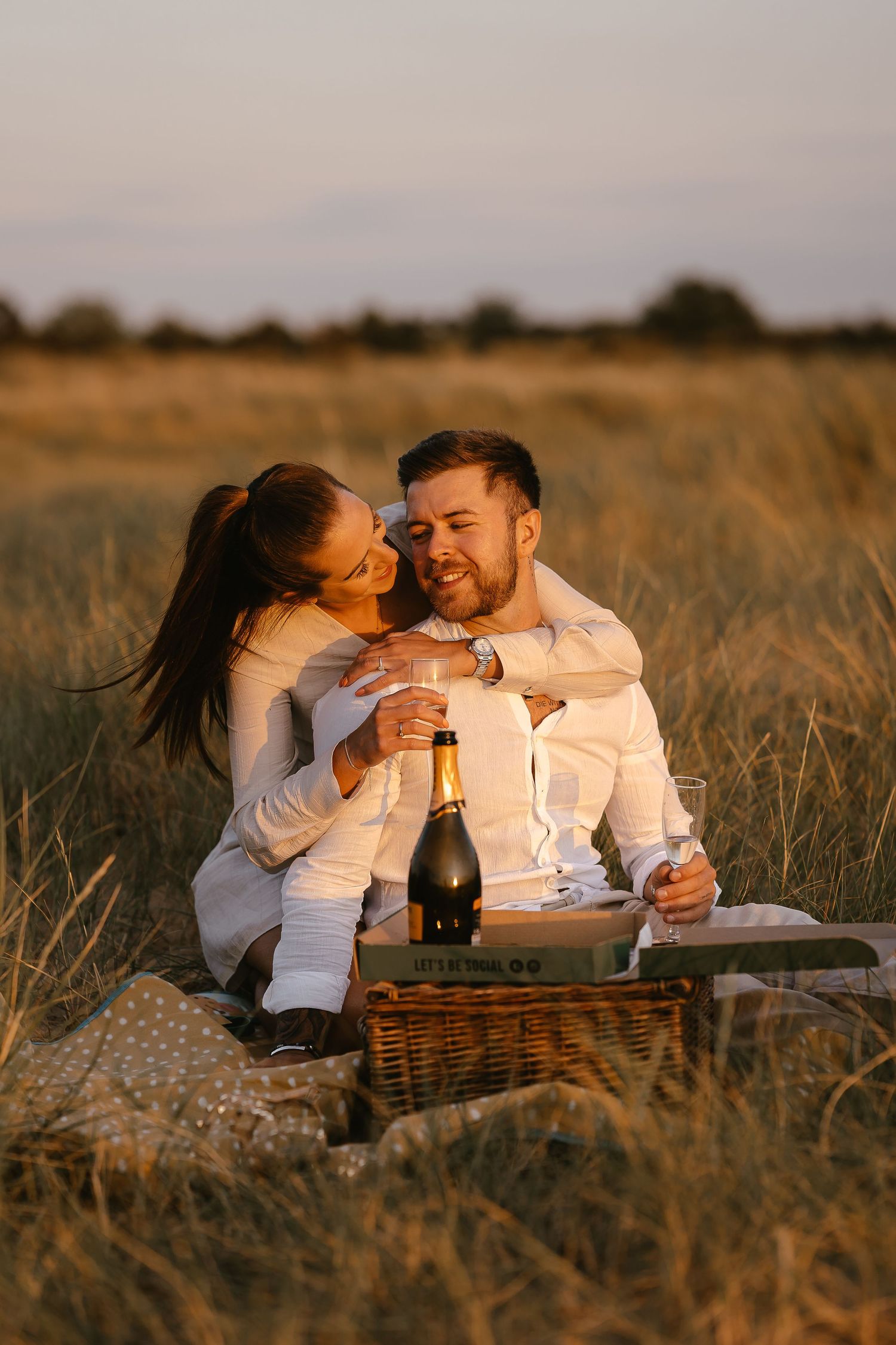 Couple embracing and laughing at a sunset picnic in a wheat field with champagne and wicker basket.