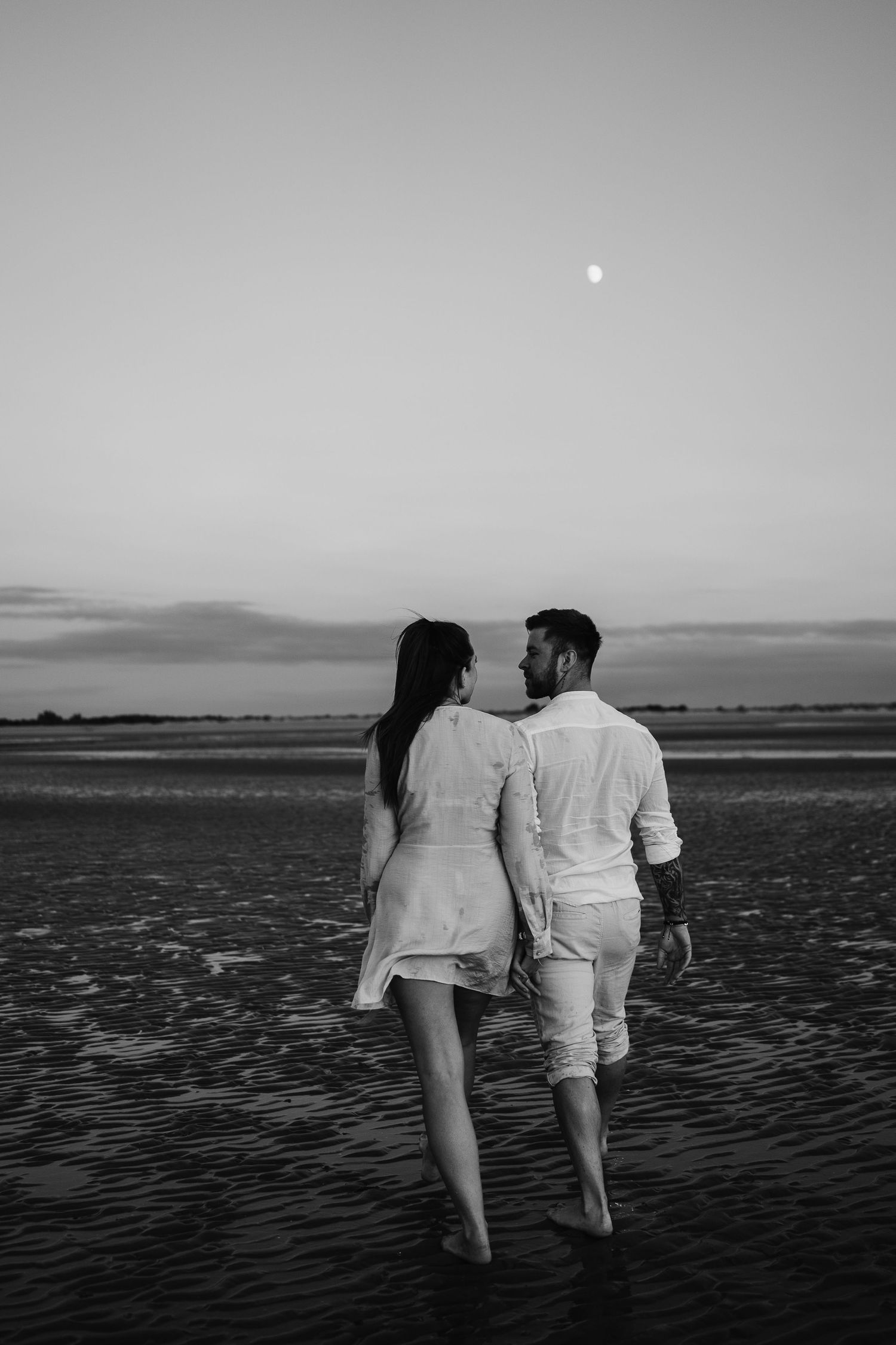 Black and white photo of couple walking hand in hand on beach under moonlit sky.