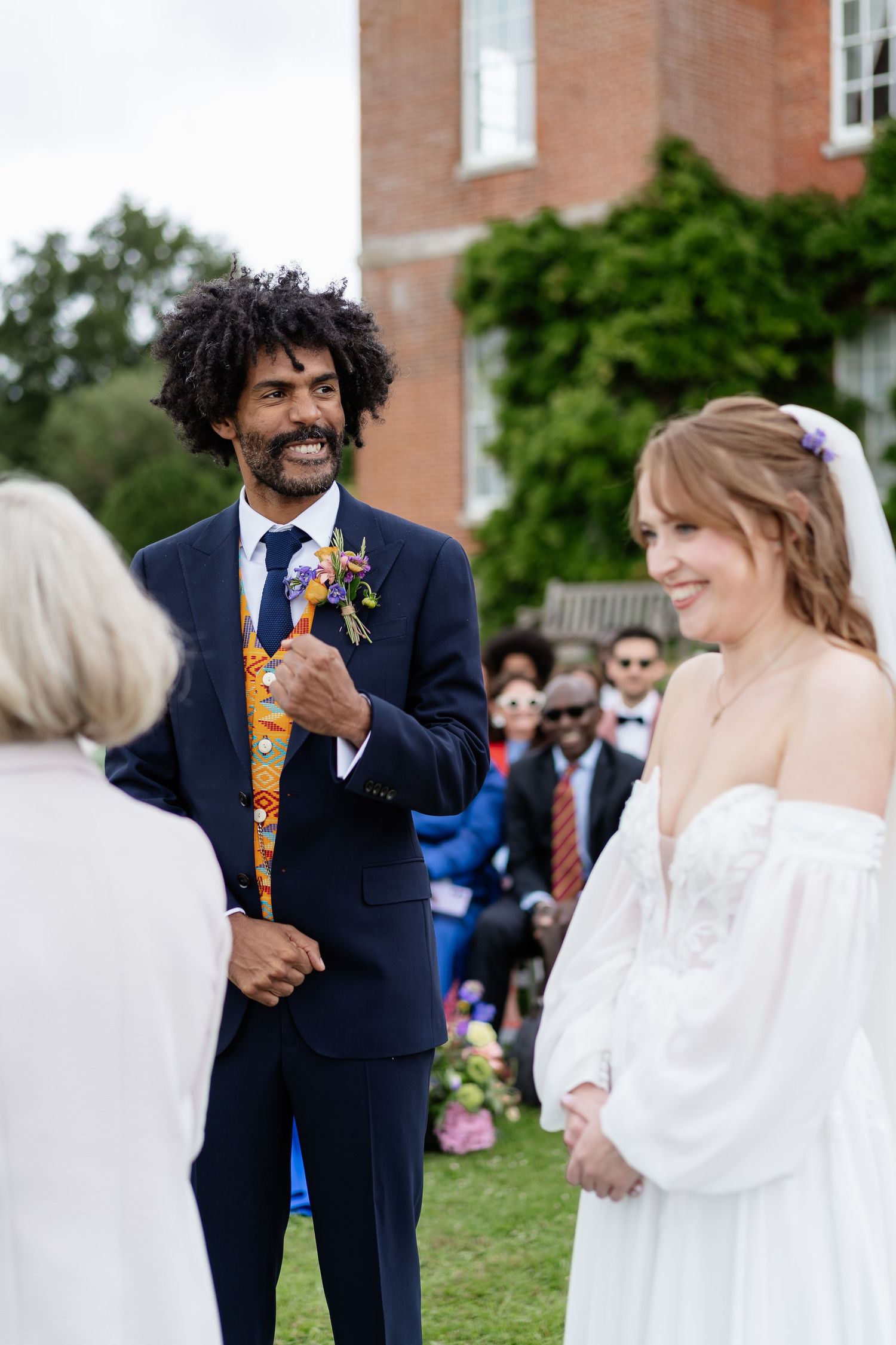 Groom in navy suit with colorful vest smiles at bride in white off-shoulder gown during outdoor wedding ceremony.
