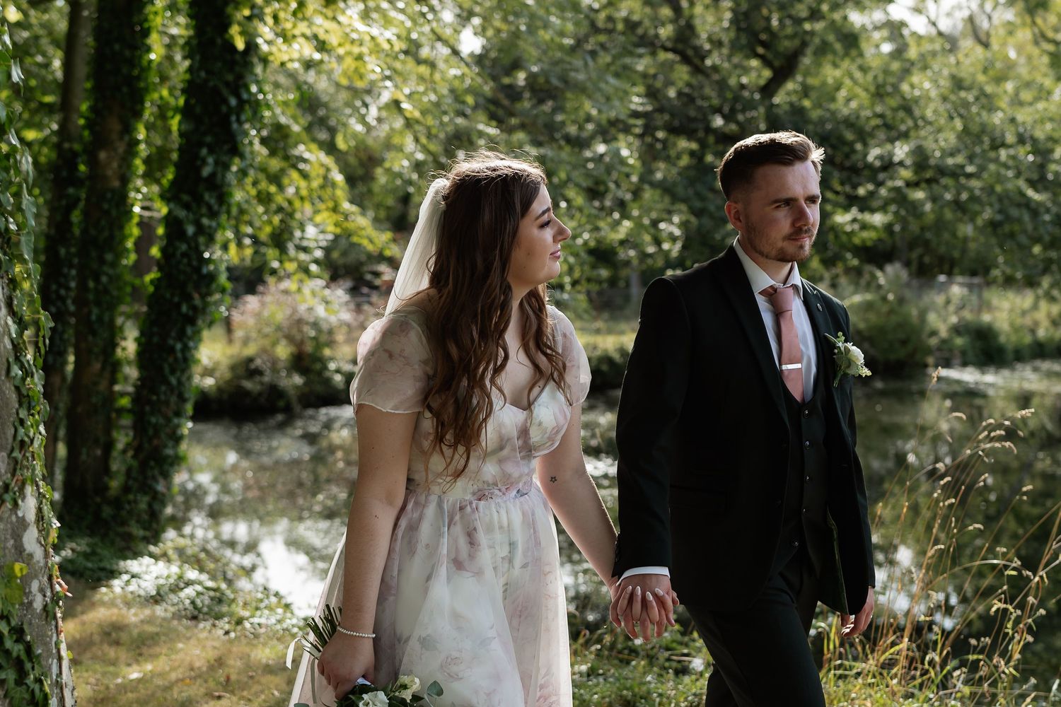 Bride and groom hold hands walking along lush green riverbank at romantic outdoor wedding venue.
