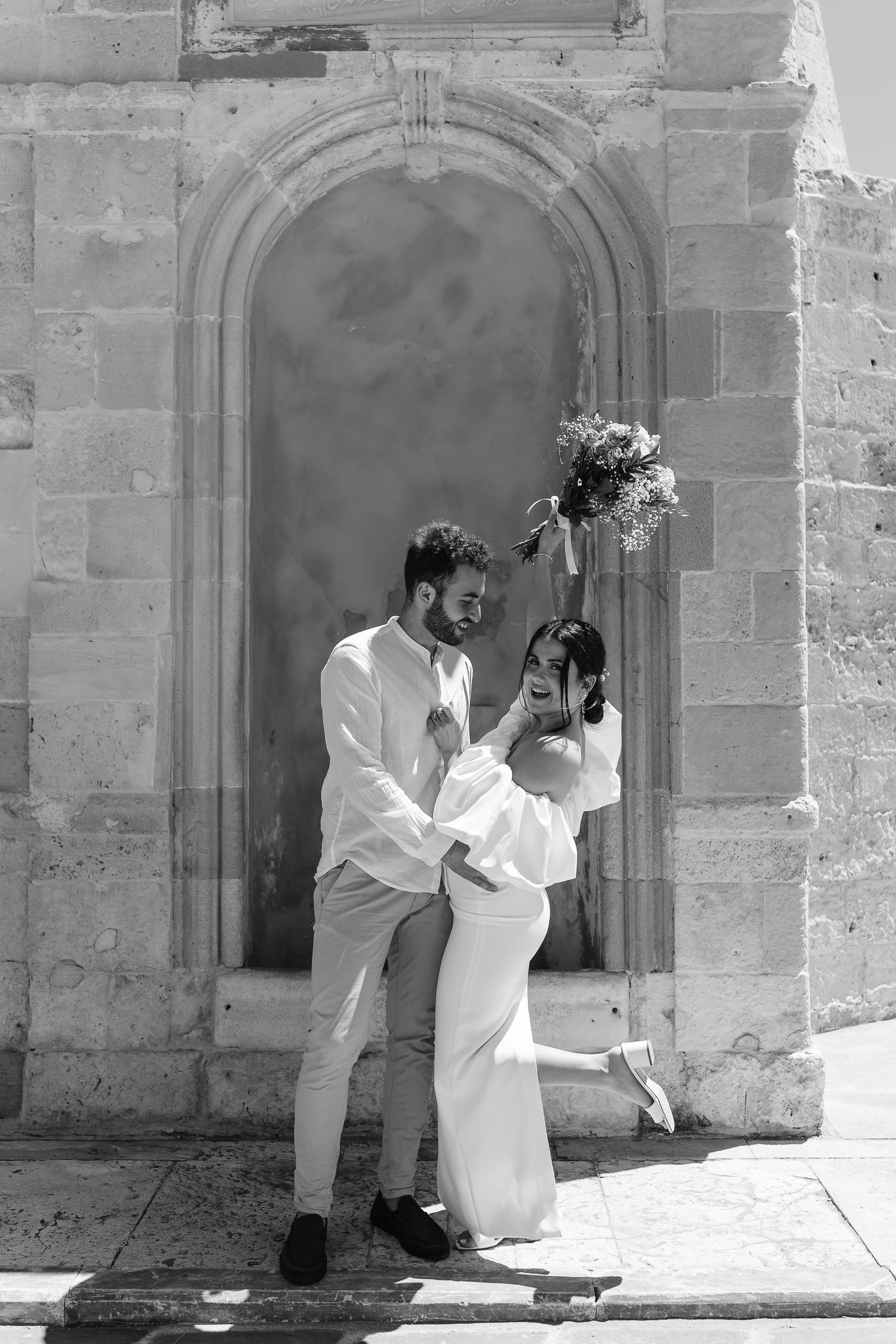 Black and white image of laughing couple embracing playfully in front of an arched stone doorway on their wedding day.