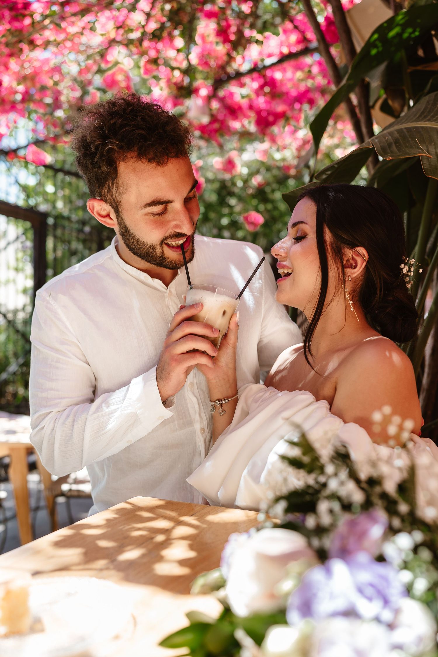 Happy couple sharing an iced drink together at an outdoor cafe beneath blooming pink bougainvillea.