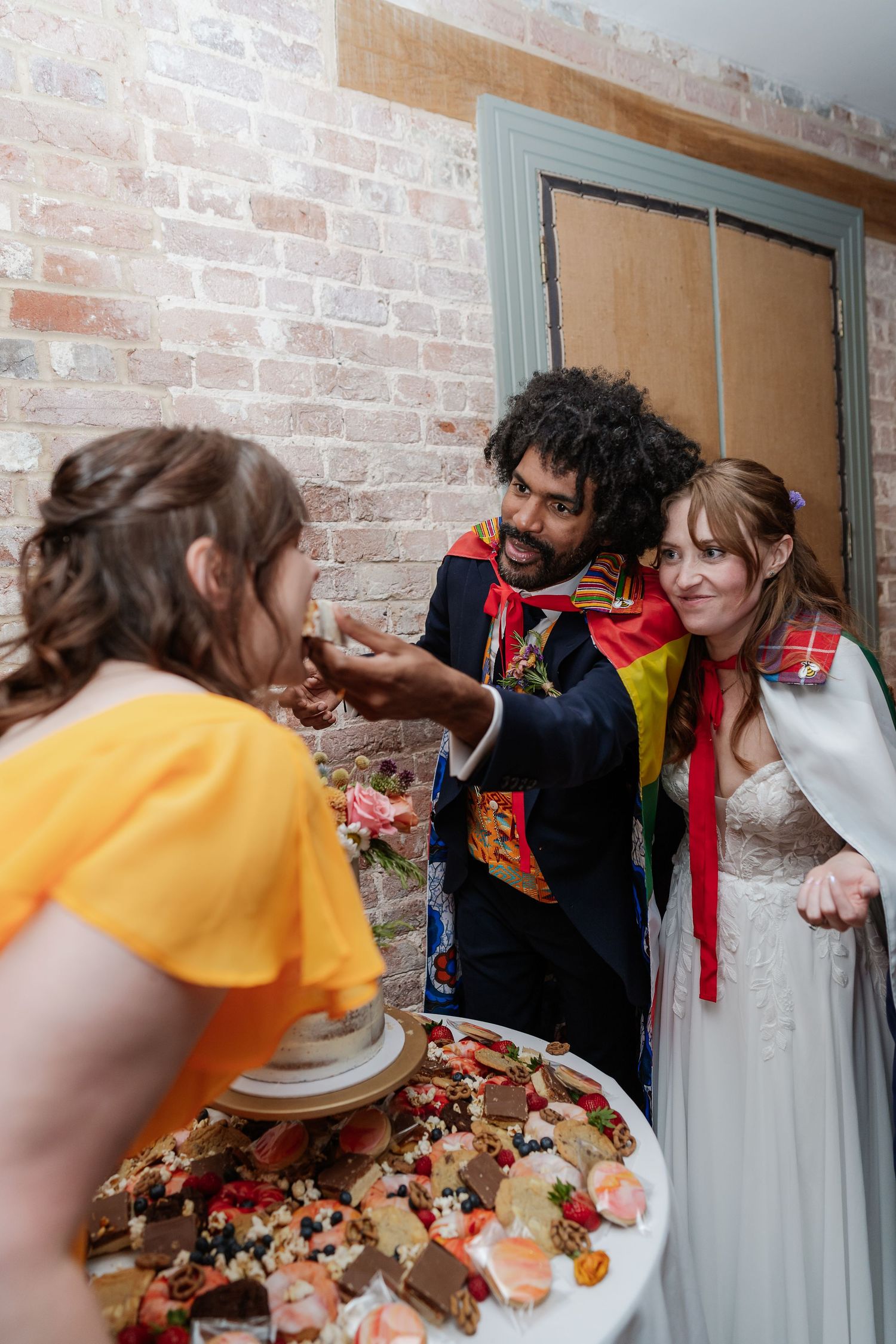 Bride and groom in colorful capes share wedding cake with guest at reception.