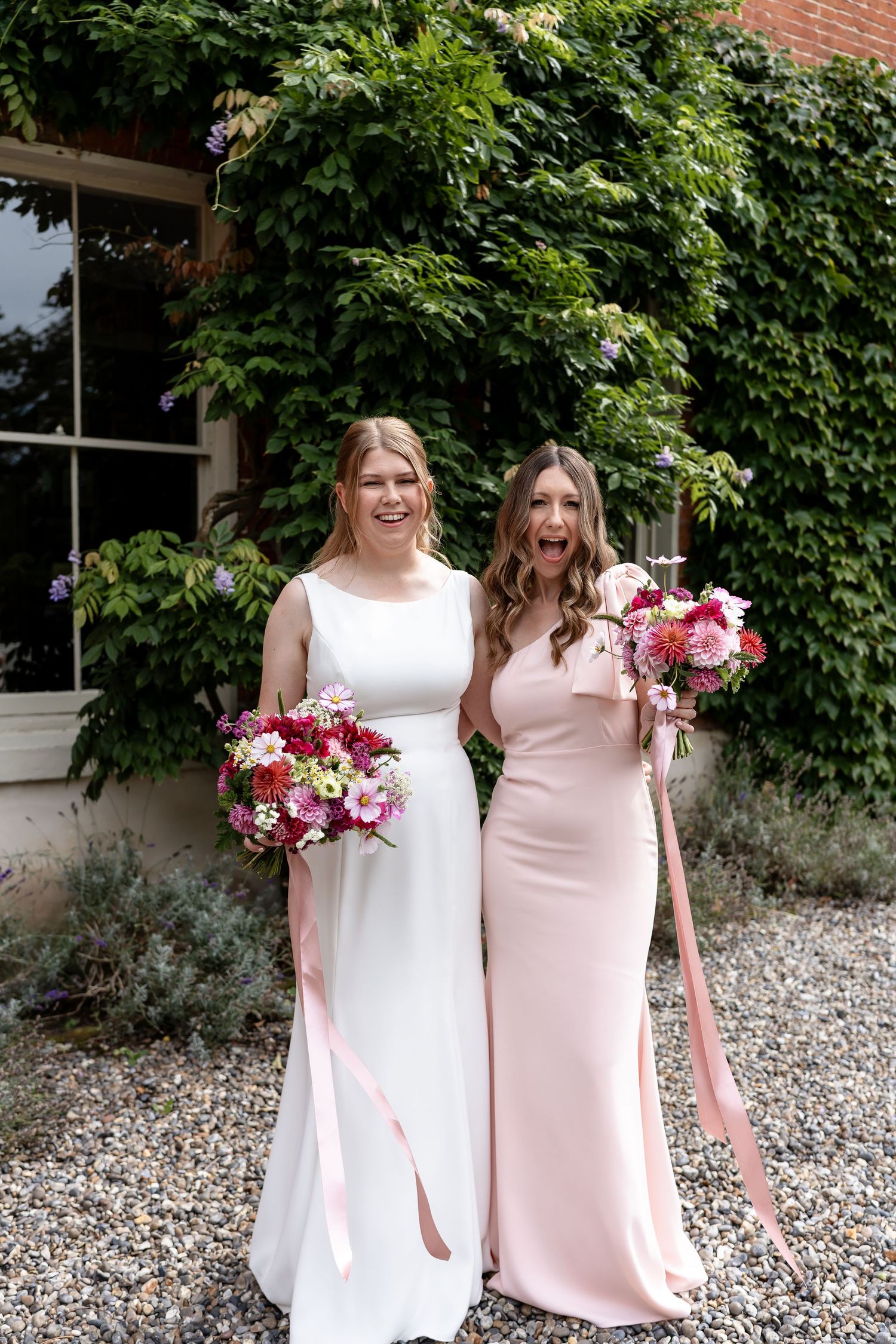 Bride in white gown and bridesmaid in blush pink dress laugh holding colorful bouquets before ivy-covered brick wall.