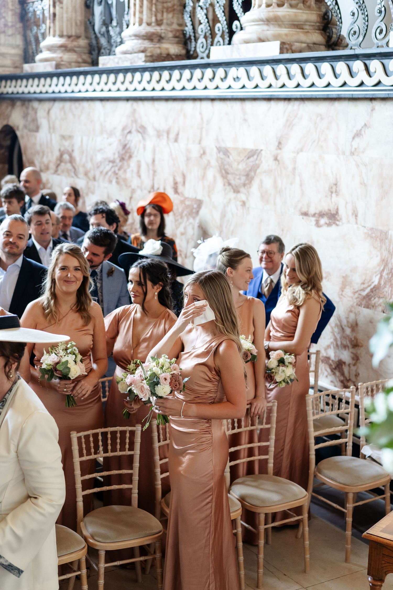 Bridesmaids in rose gold satin gowns holding floral bouquets stand in a grand marble wedding venue aisle.