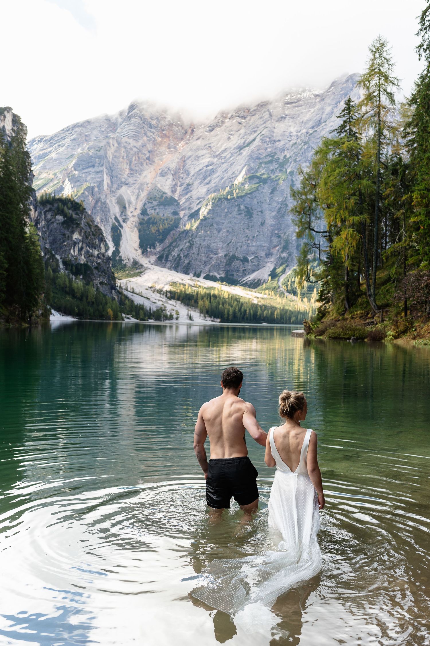 Adventurous couple wading in crystal clear alpine lake surrounded by dramatic Dolomite mountain scenery.